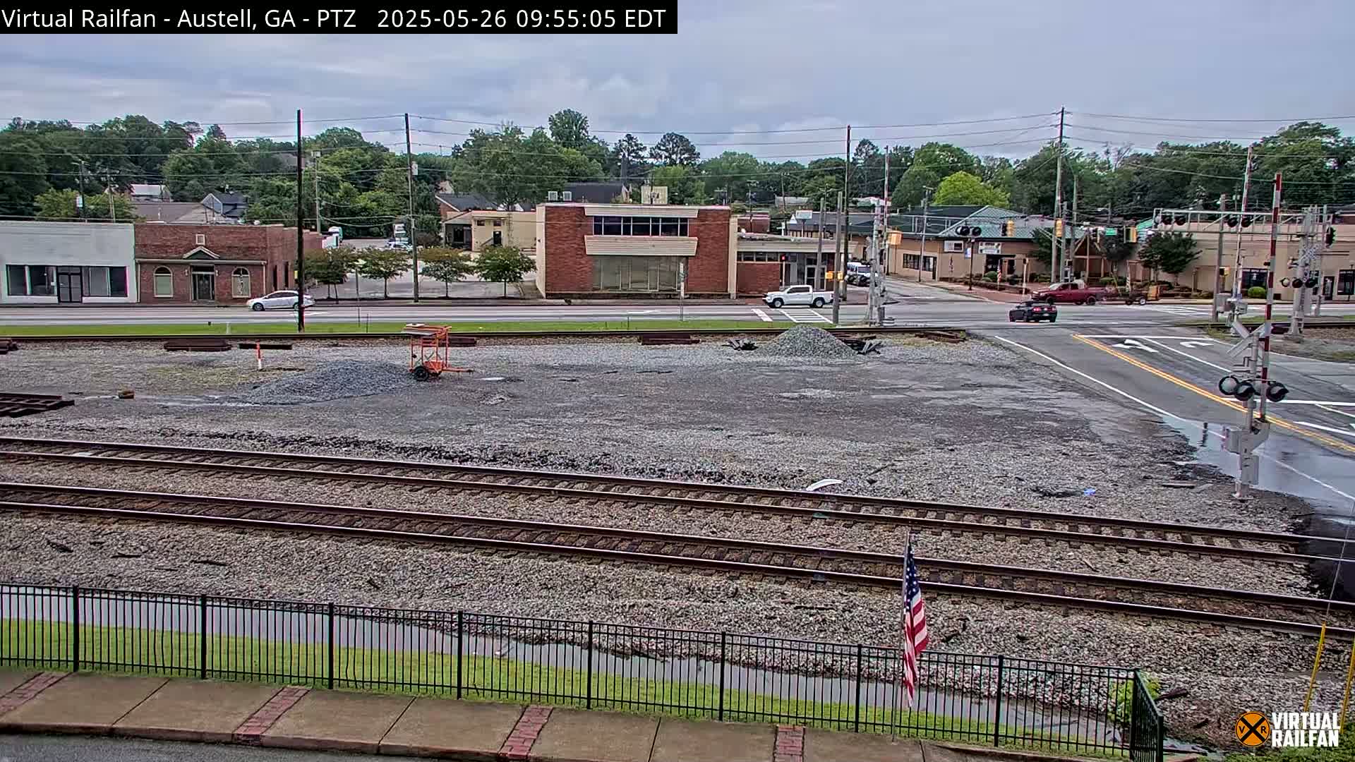 A view of railroad tracks and a gravel area next to a street with buildings and cars, under an overcast sky with some wet patches visible.