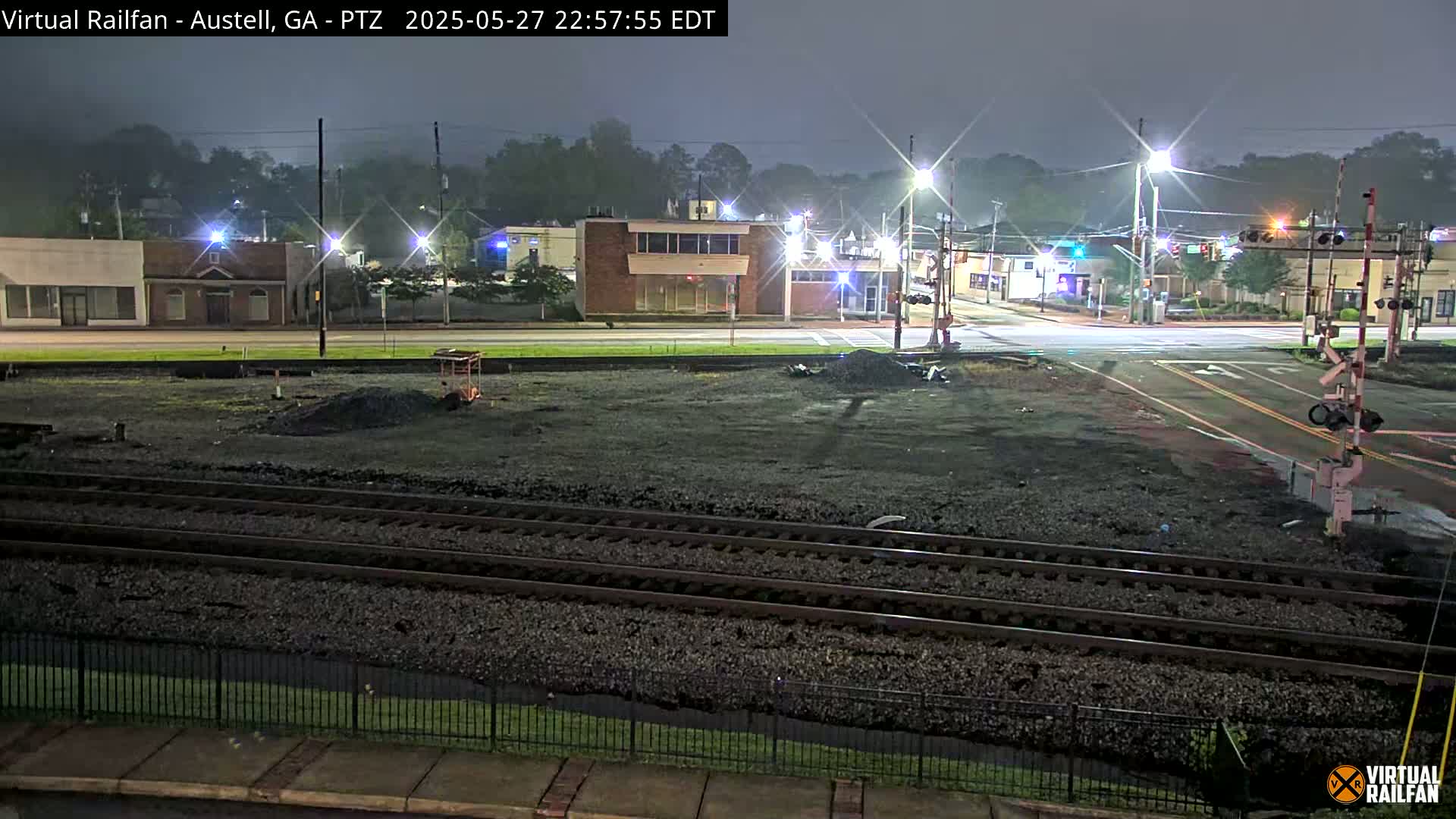 A nighttime view of railroad tracks intersecting with a street in front of buildings under hazy conditions.