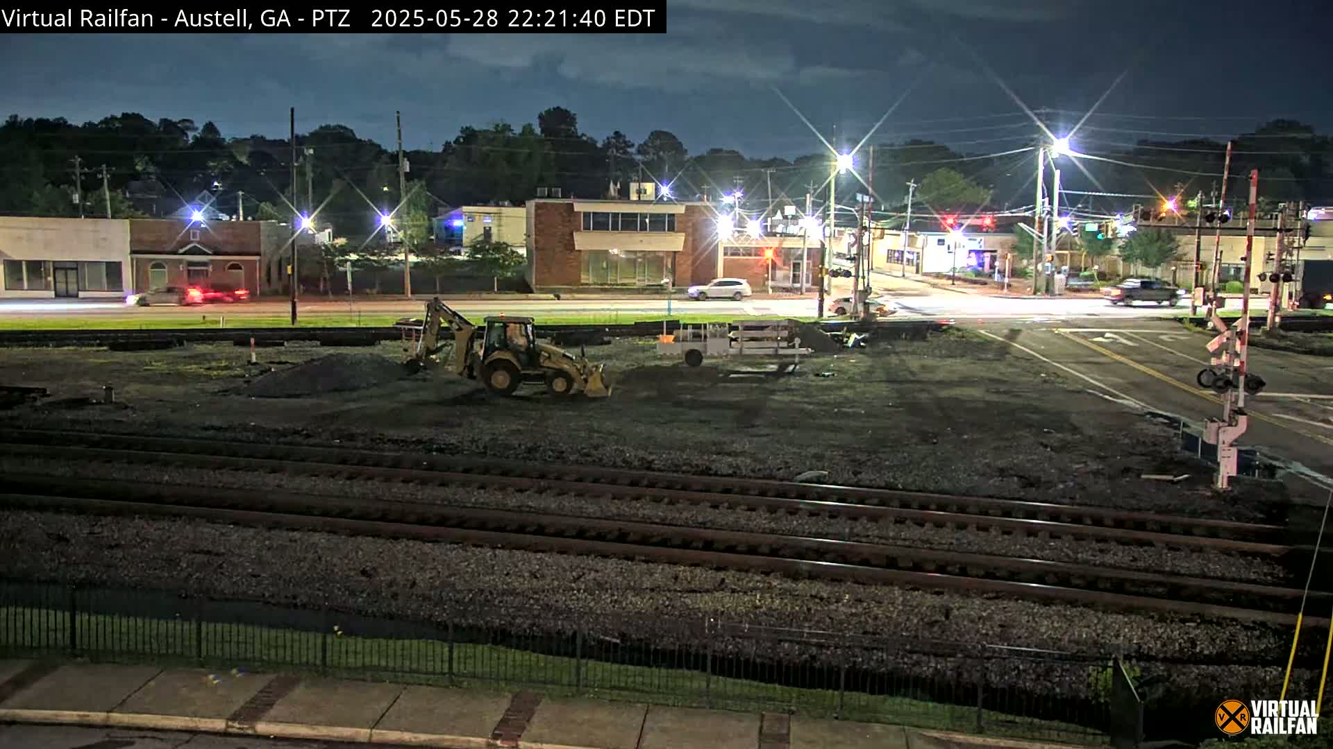 At night, under a clear sky, construction equipment sits on a plot of land next to railroad tracks near a street intersection with cars and buildings in the background.