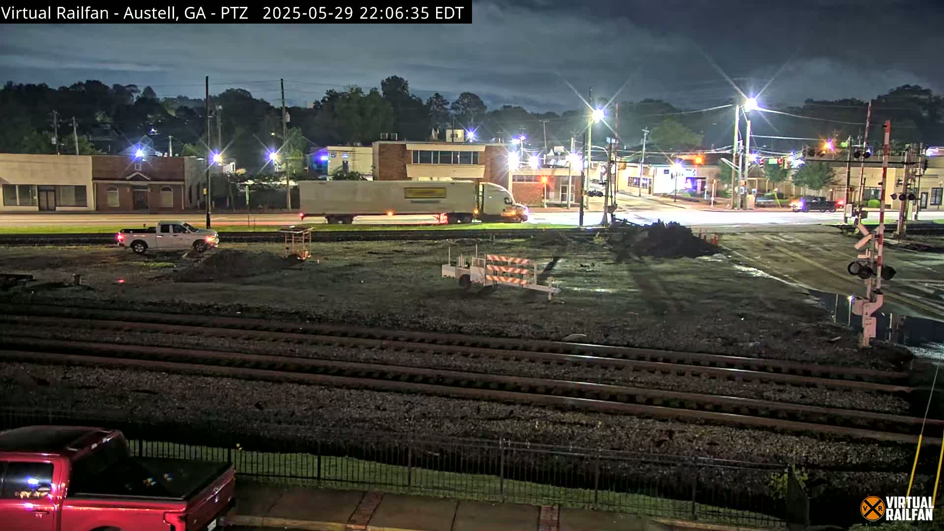 A nighttime view of a railway crossing with construction in progress, featuring several vehicles, streetlights illuminating the scene under a partly cloudy sky.