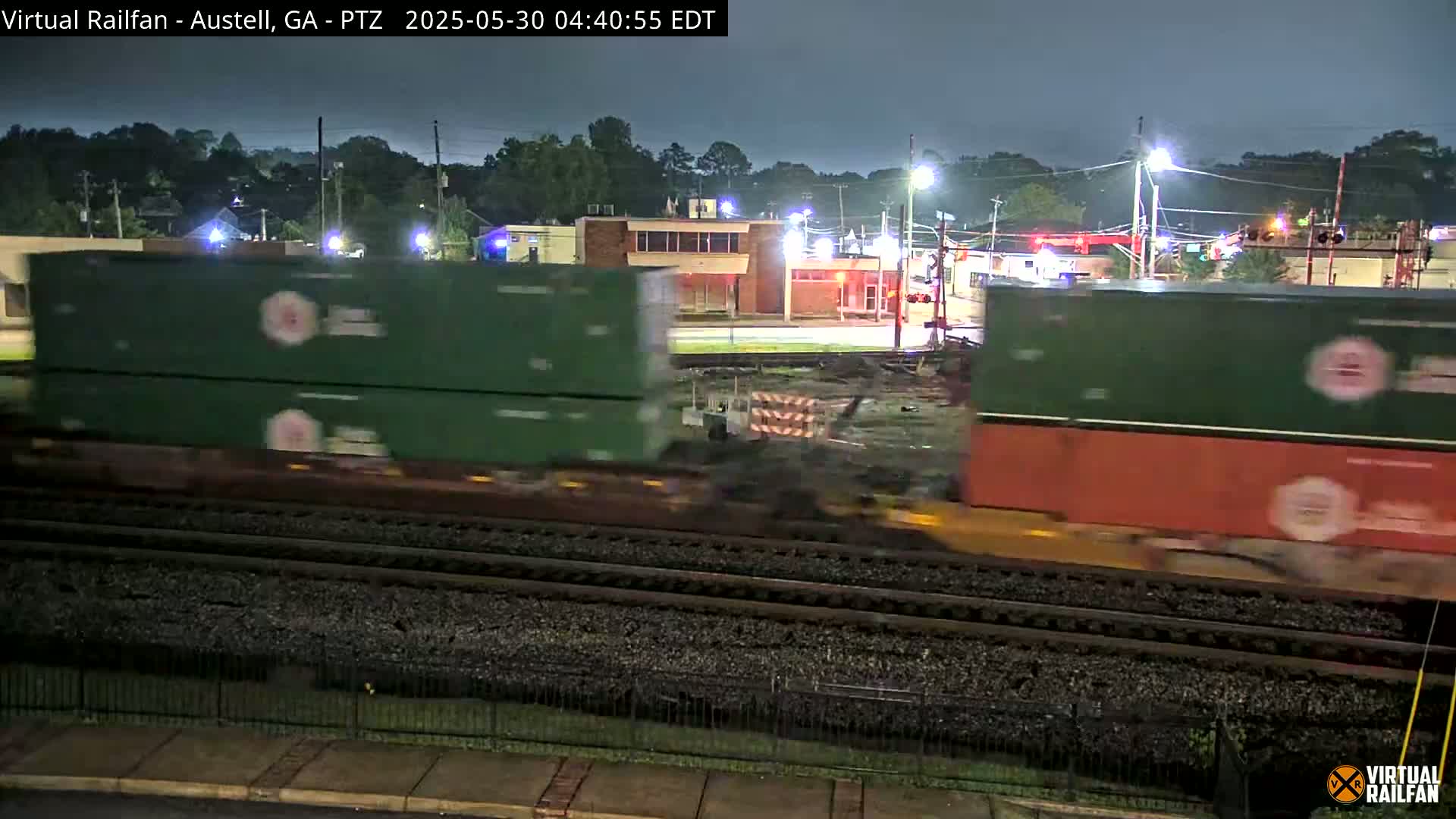 A nighttime view of a freight train moving past a building and crossing under streetlights in the dark.