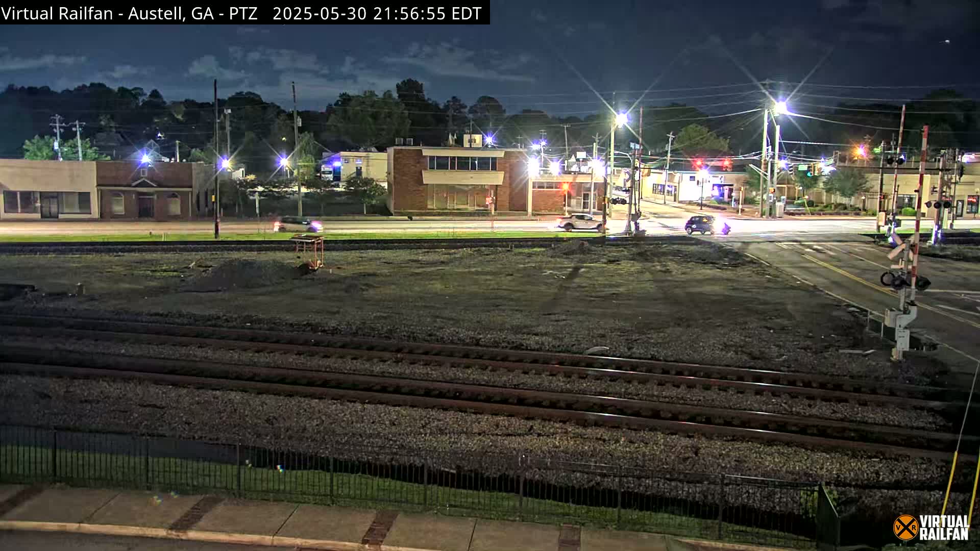 A nighttime view of a railroad crossing near a street intersection with several buildings and sparse vehicle traffic under a partly cloudy sky.