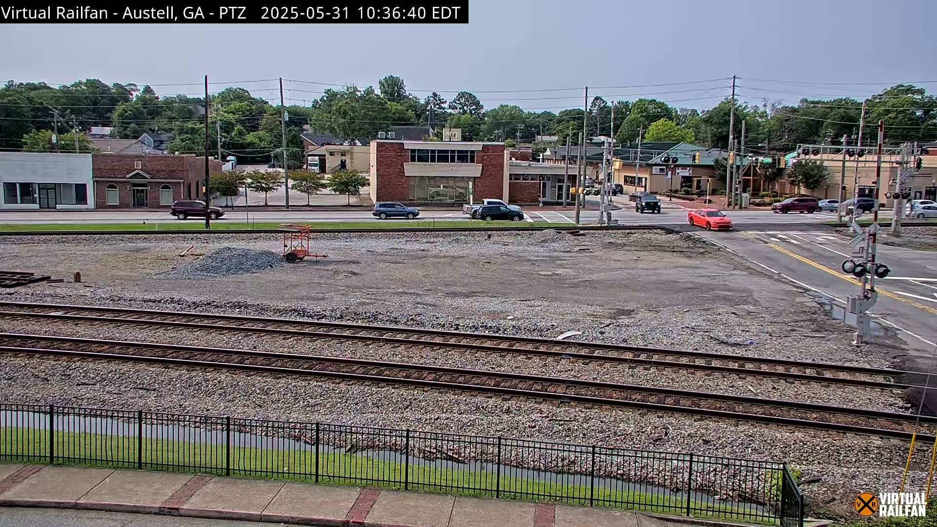 A mostly sunny view of a gravel lot with train tracks and a black metal fence in front of a street intersection with several cars and buildings in the background.