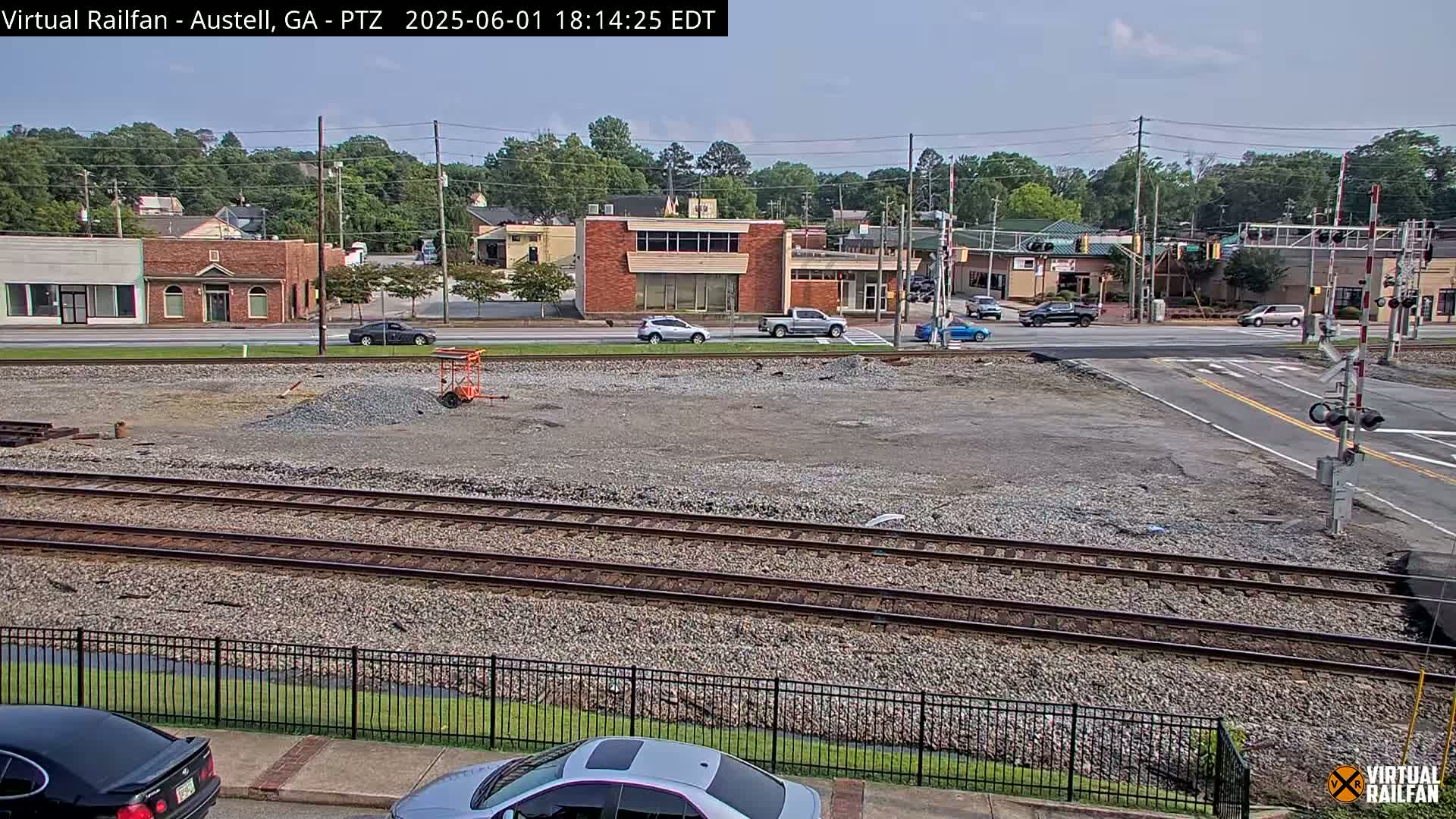 A mostly sunny day reveals a gravel lot next to railroad tracks and a street intersection, with several cars and buildings visible in the background.