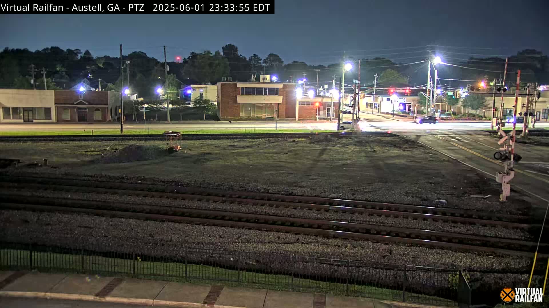 A nighttime view of a railroad crossing near a street intersection with several low-rise buildings in the background under a clear, dark sky.