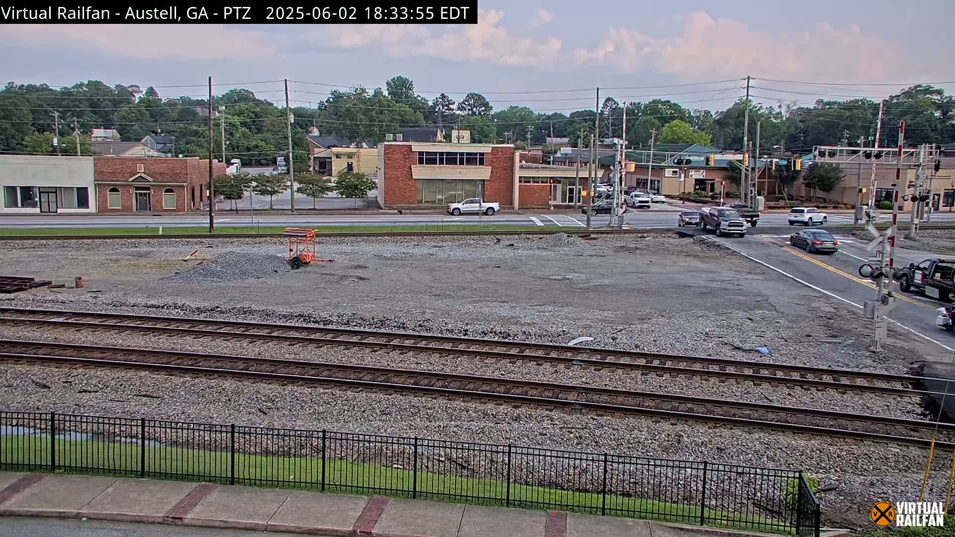 A mostly clear, overcast day reveals a view of railroad tracks beside a roadway with several buildings and vehicles, and a large empty gravel lot in the foreground.