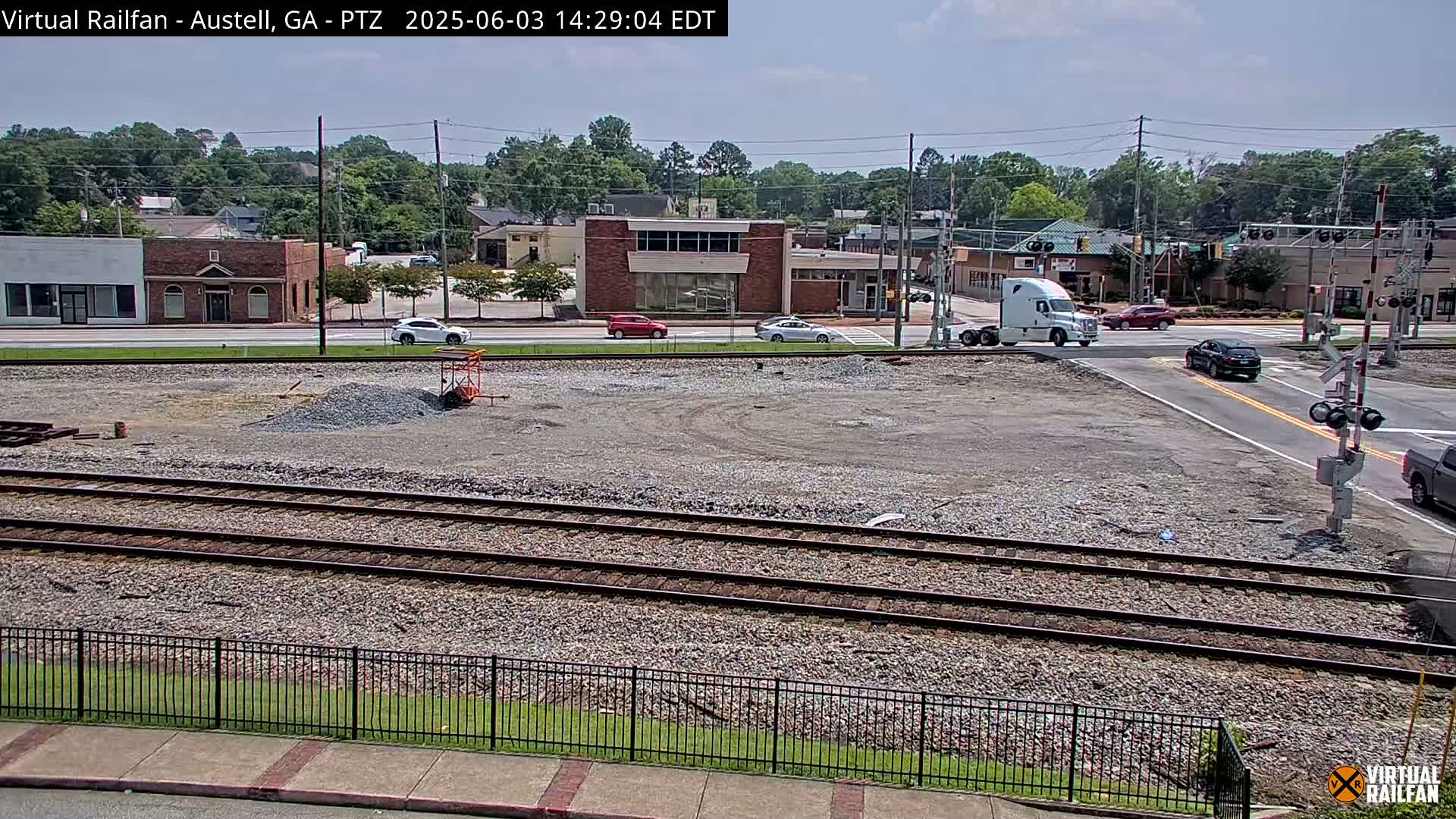 A mostly sunny view of a railway crossing in a town, with a gravel area under construction and several vehicles driving on the road.