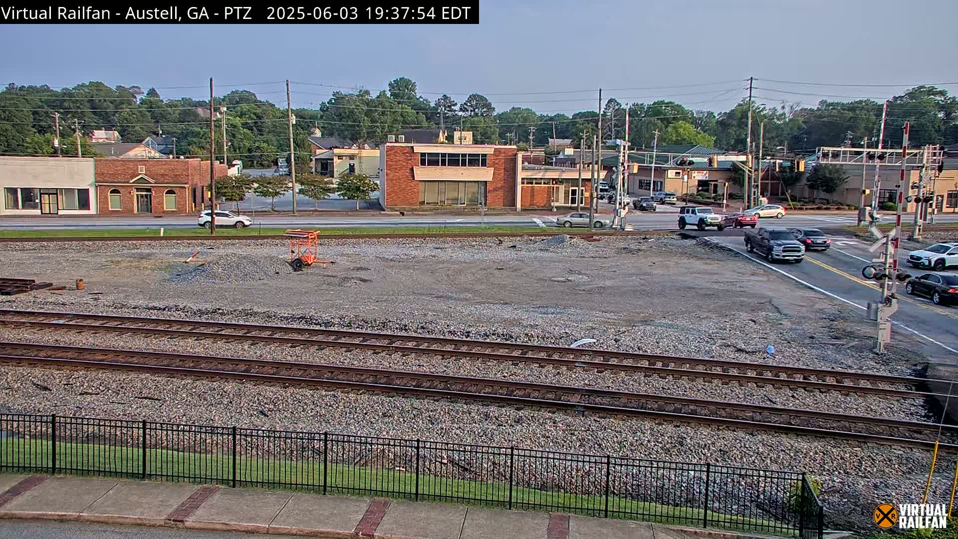 A mostly clear day shows a view of railroad tracks next to a mostly empty lot, bordered by a street with buildings and cars, some of which are at a nearby intersection.