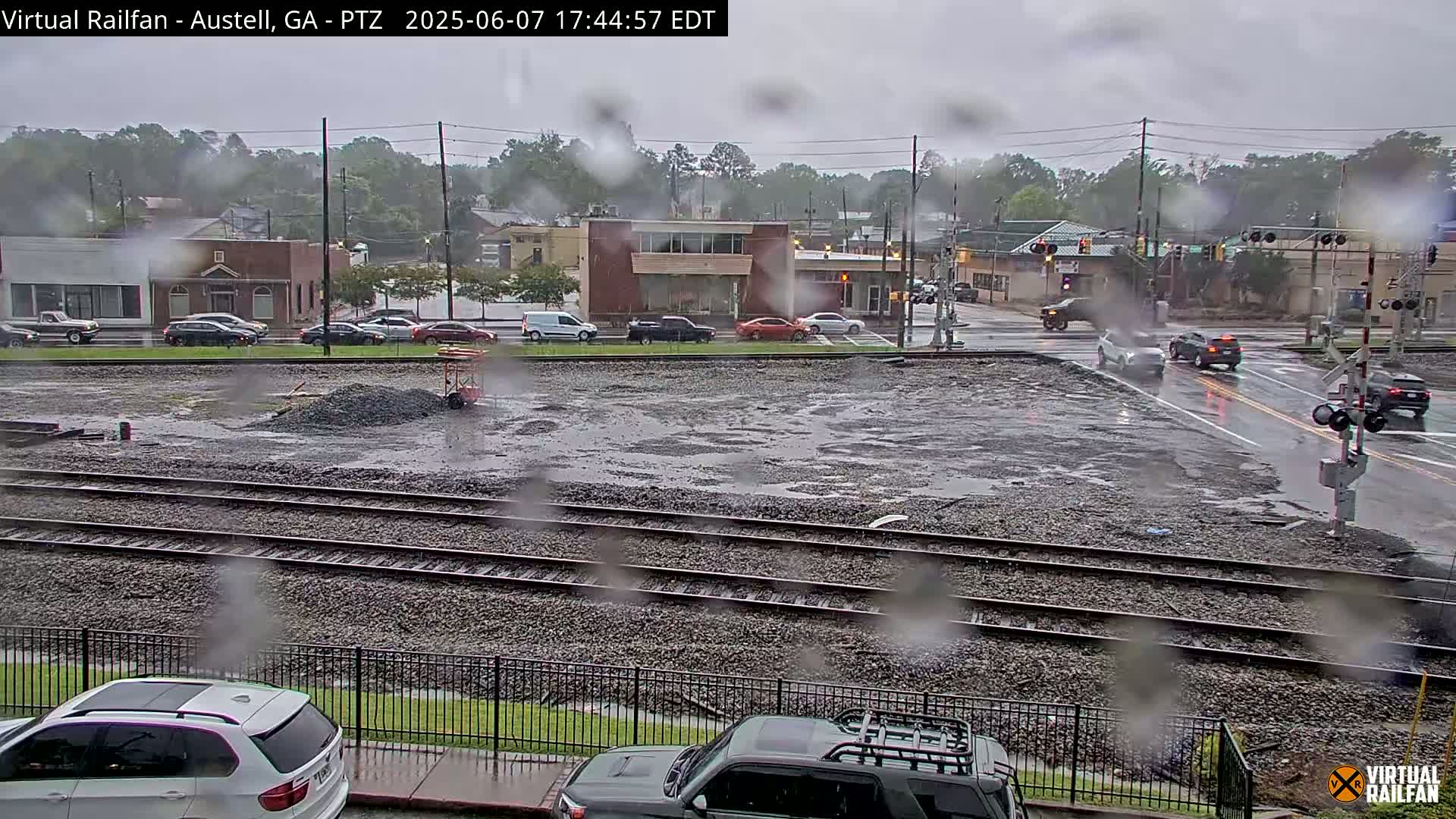 A rain-soaked view of a railroad crossing with several cars at an intersection, a pile of gravel, and buildings in the background.