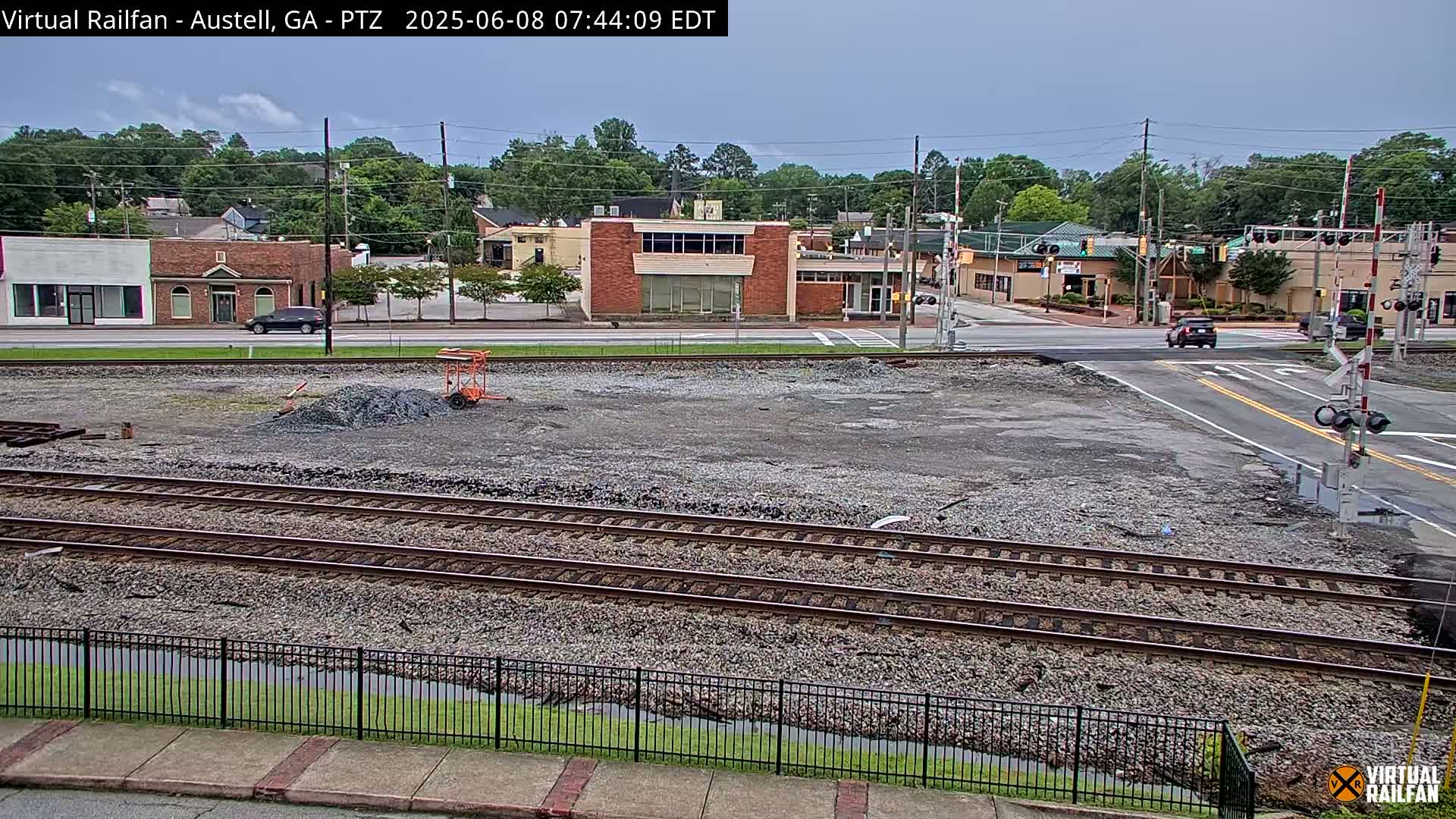 A mostly empty, gravel-covered lot with train tracks sits beside a street with buildings and cars under a partly cloudy sky.