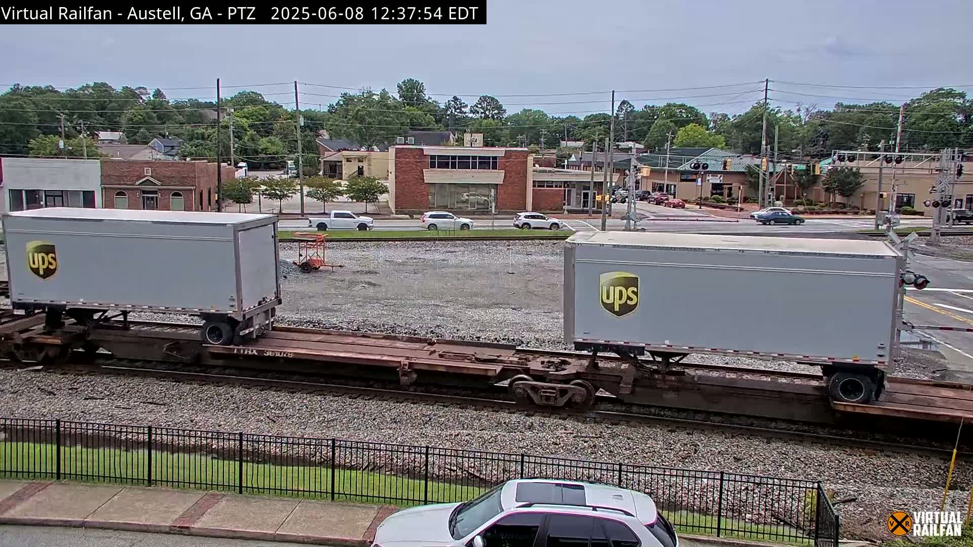 Two UPS trailers are on a flatbed railcar, next to a street with cars and buildings under a partly cloudy sky.