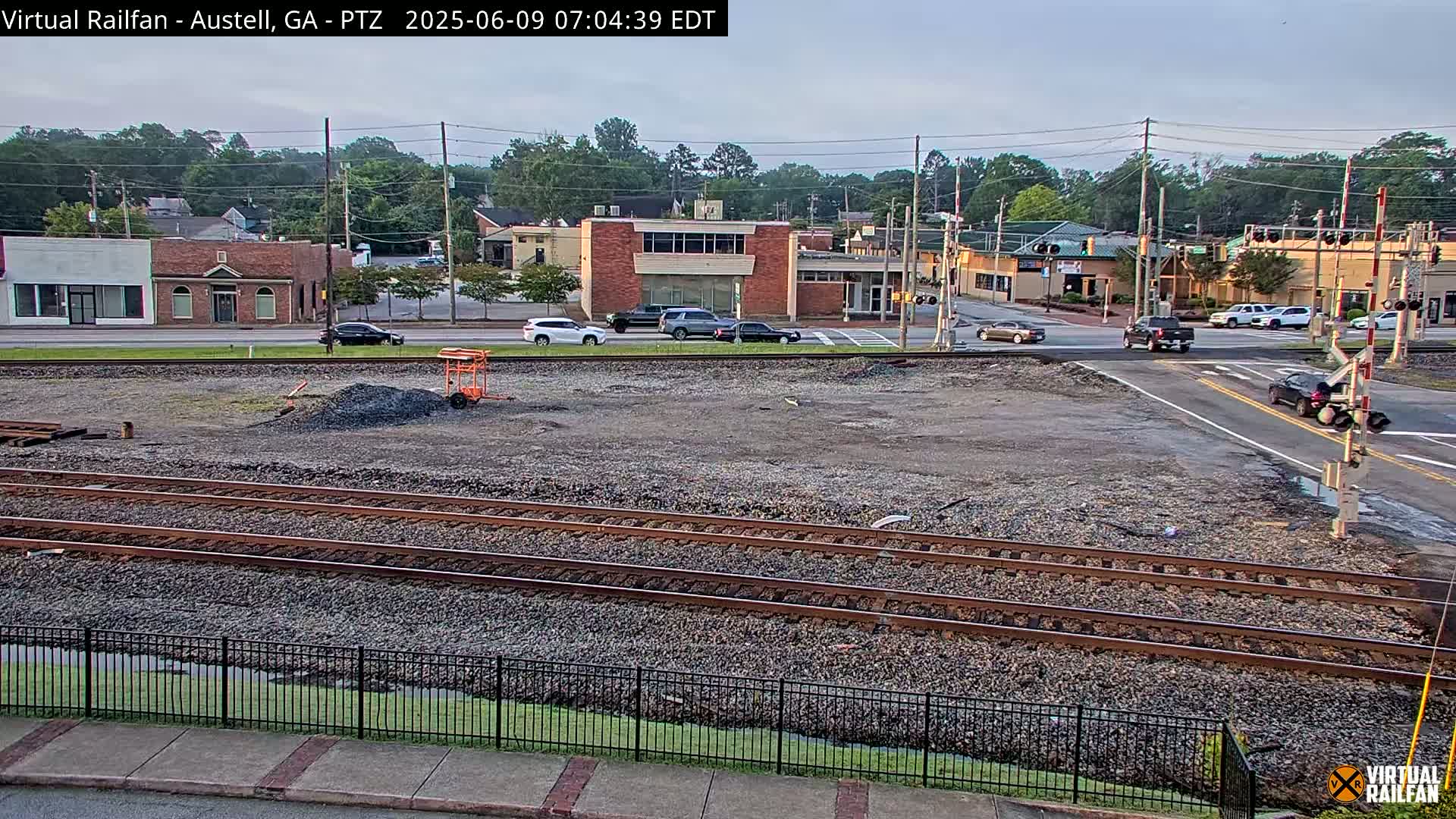 A view of railroad tracks and a street intersection in a town, with several cars visible and a mostly clear sky.