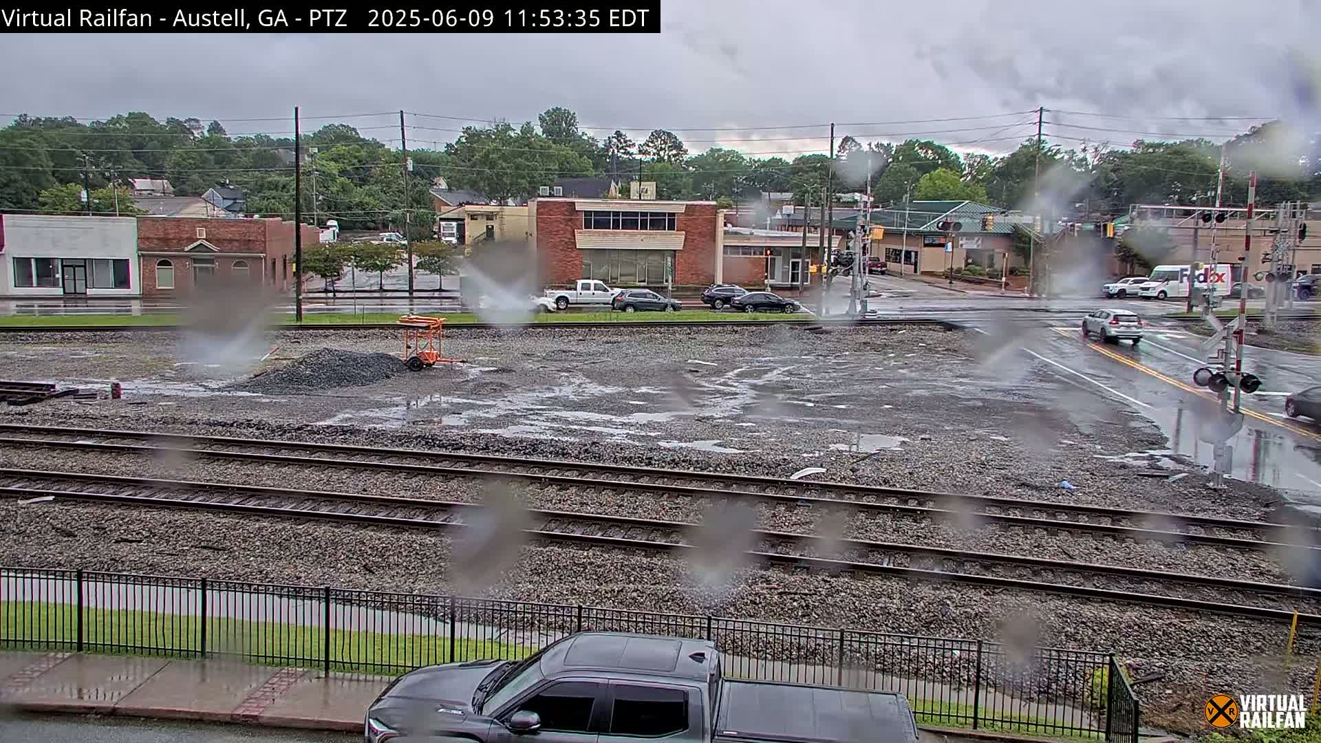 A rain-soaked, gravel-covered area with train tracks sits in front of a street and buildings under a cloudy sky.