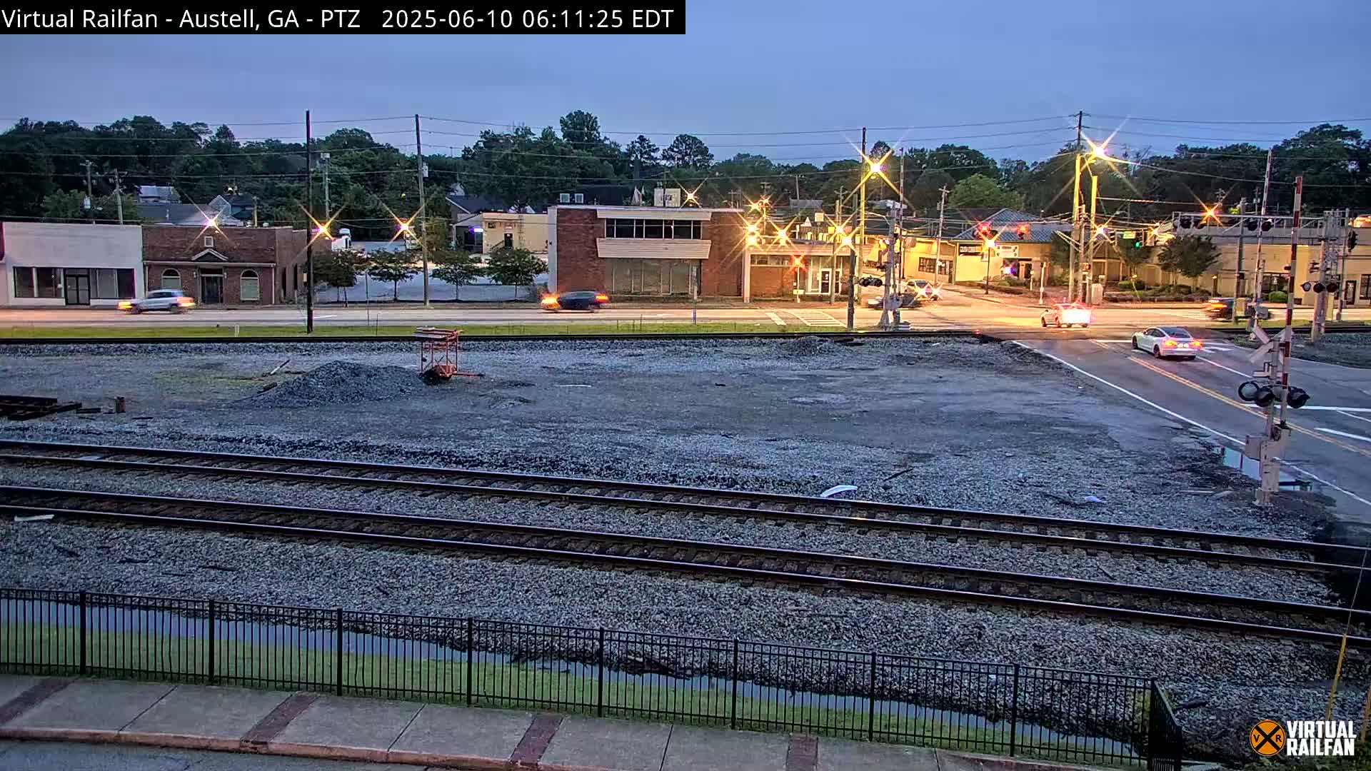A twilight view of a railroad crossing near a street intersection in a town, with several cars visible and a pile of gravel in the foreground.