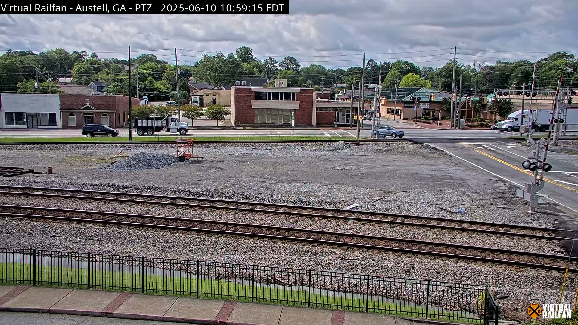 A mostly cloudy day shows a view of railroad tracks and a road intersection near some low-rise buildings, with a gravel lot in the foreground.
