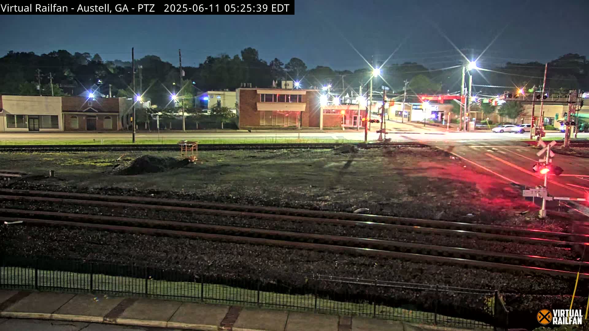 A nighttime view of a railroad crossing with several buildings and streetlights visible under a clear sky.