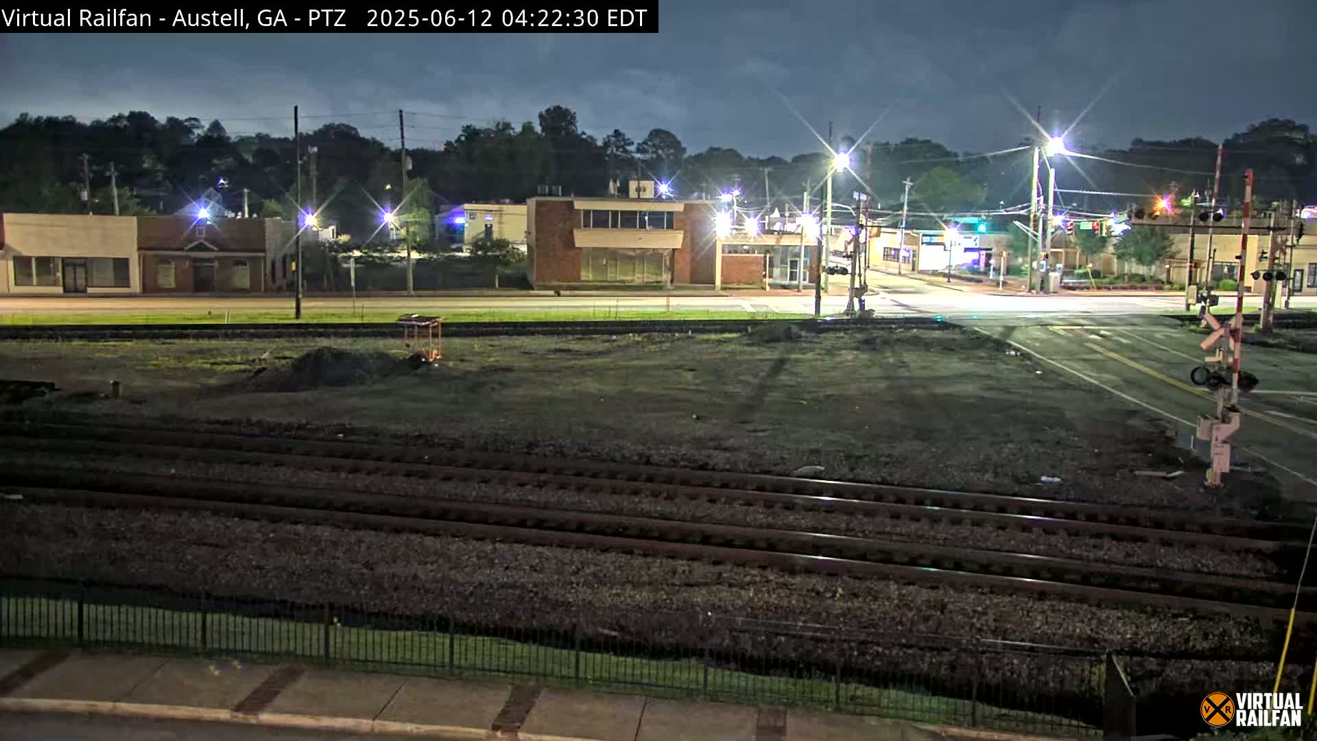 A nighttime view of a street intersection with train tracks in the foreground, under a partly cloudy sky.