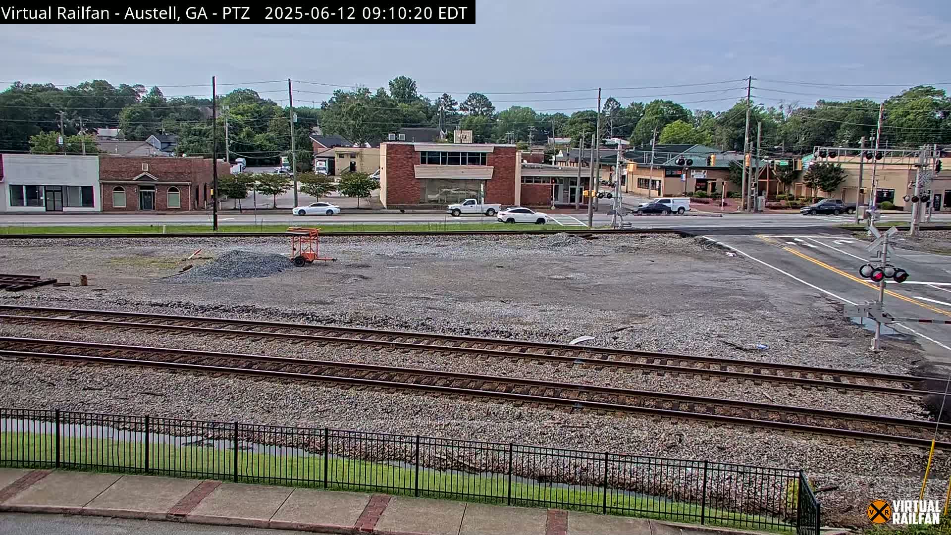 A mostly cloudy day shows a view of a railway crossing in front of a street with several buildings, a small pile of gravel, and a few cars.