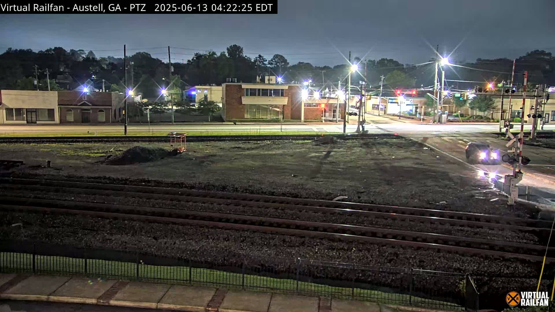 At night, a dark, empty lot with train tracks sits between a street intersection with a car and several low buildings under streetlights.