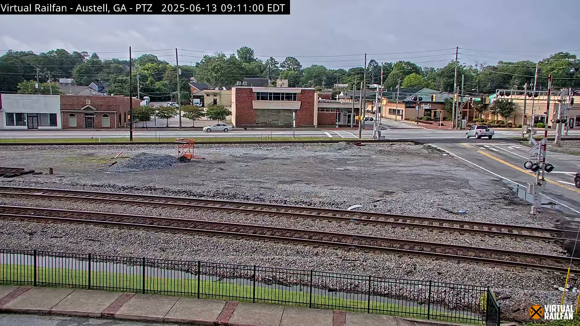 A mostly clear day reveals railroad tracks intersecting a street in front of several commercial buildings; a small construction or demolition site sits in the foreground.