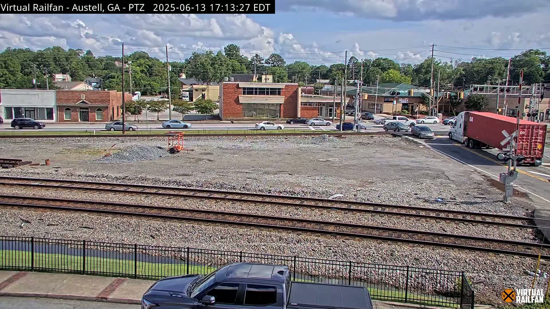 A mostly sunny day shows a section of train tracks in a town with several cars on the road and a red container truck at a crossing.