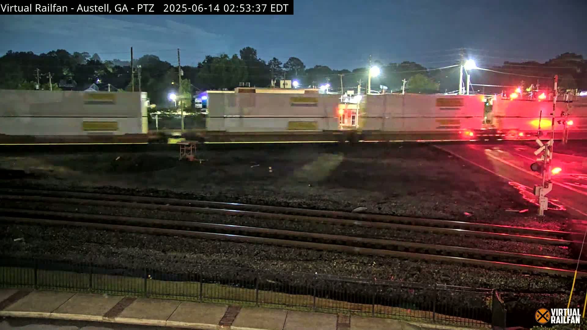 A mostly sunny day shows a section of train tracks in a town with several cars on the road and a red container truck at a crossing.