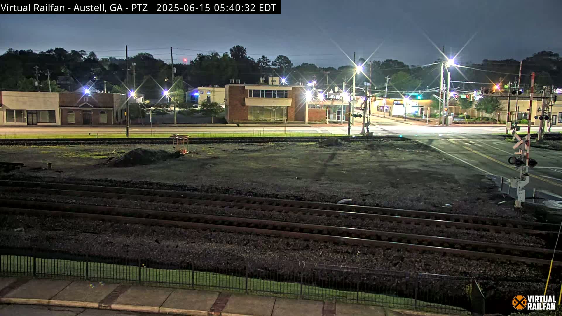A nighttime view of a train crossing in front of several low-rise buildings, under a dark, mostly clear sky.