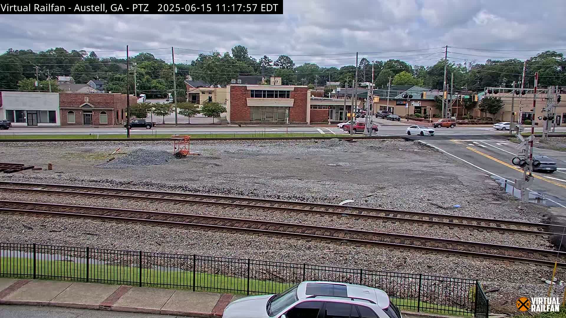 A mostly cloudy day shows a view of railroad tracks and a street intersection in front of a few buildings, with some cars visible on the street.