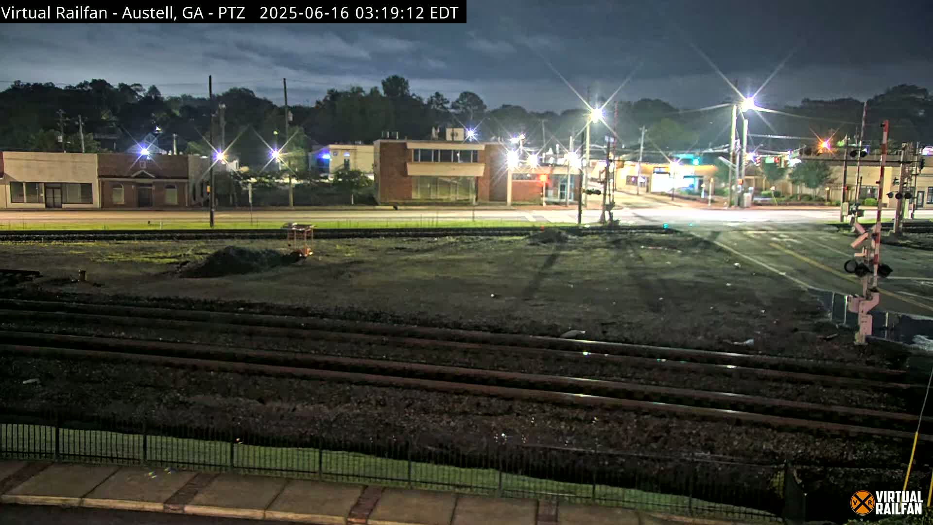 A nighttime view of a street intersection with railroad tracks in the foreground, under a mostly clear sky.