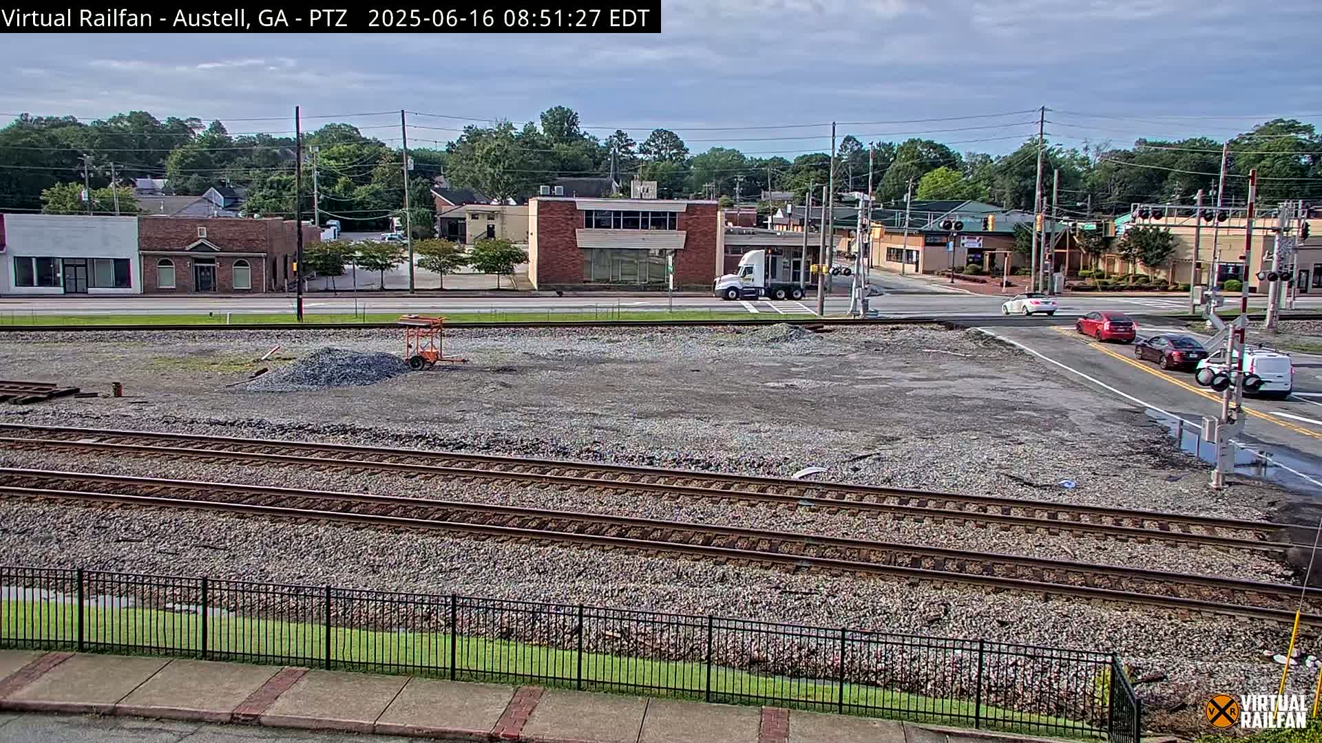 A mostly clear day view of train tracks next to a street intersection with several cars and buildings in the background.