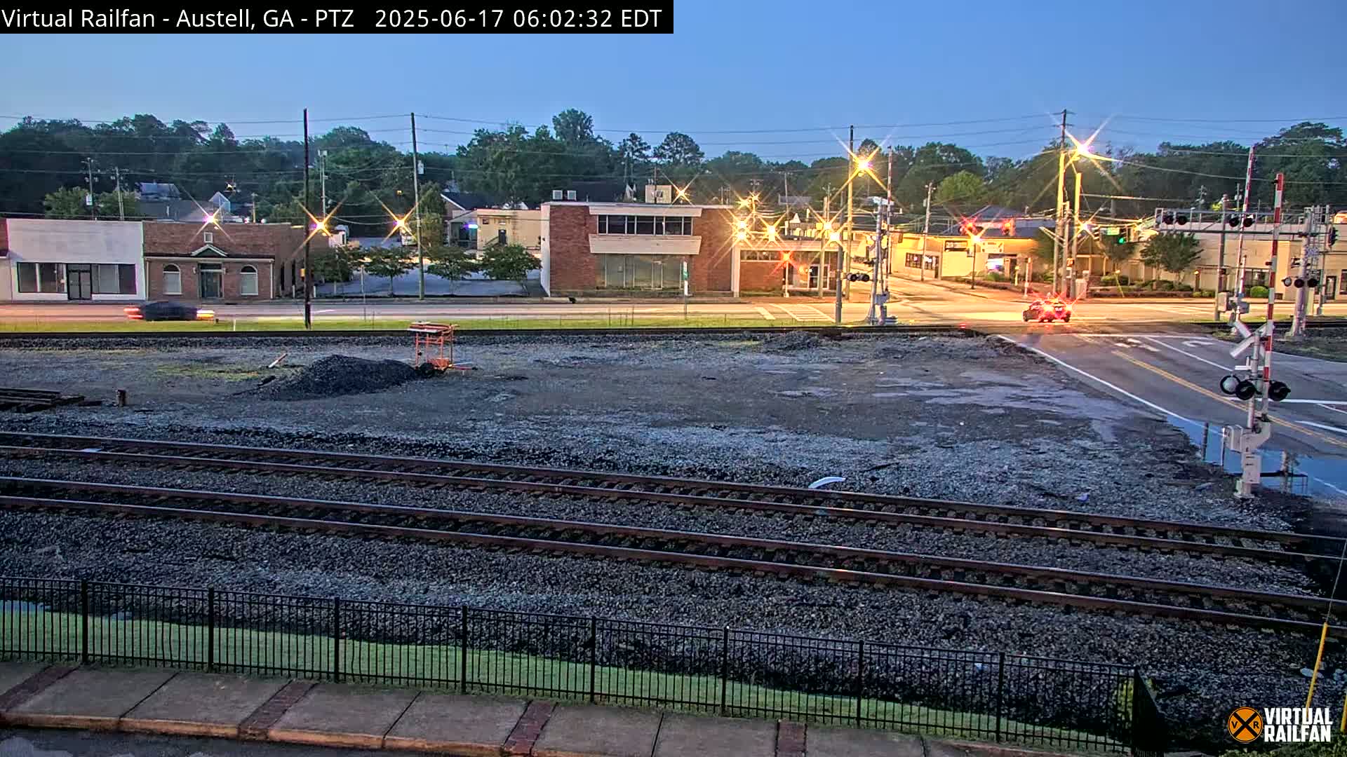 A nighttime view of a train crossing near a street with buildings, under a twilight sky.