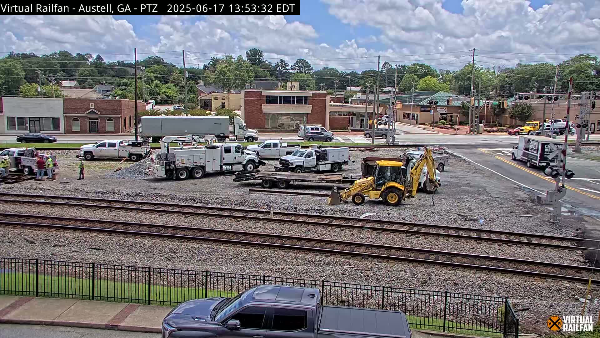 A partly cloudy day shows a construction or maintenance site near railroad tracks, with several work trucks, a backhoe, and workers present, alongside a street intersection with buildings and vehicles in the background.