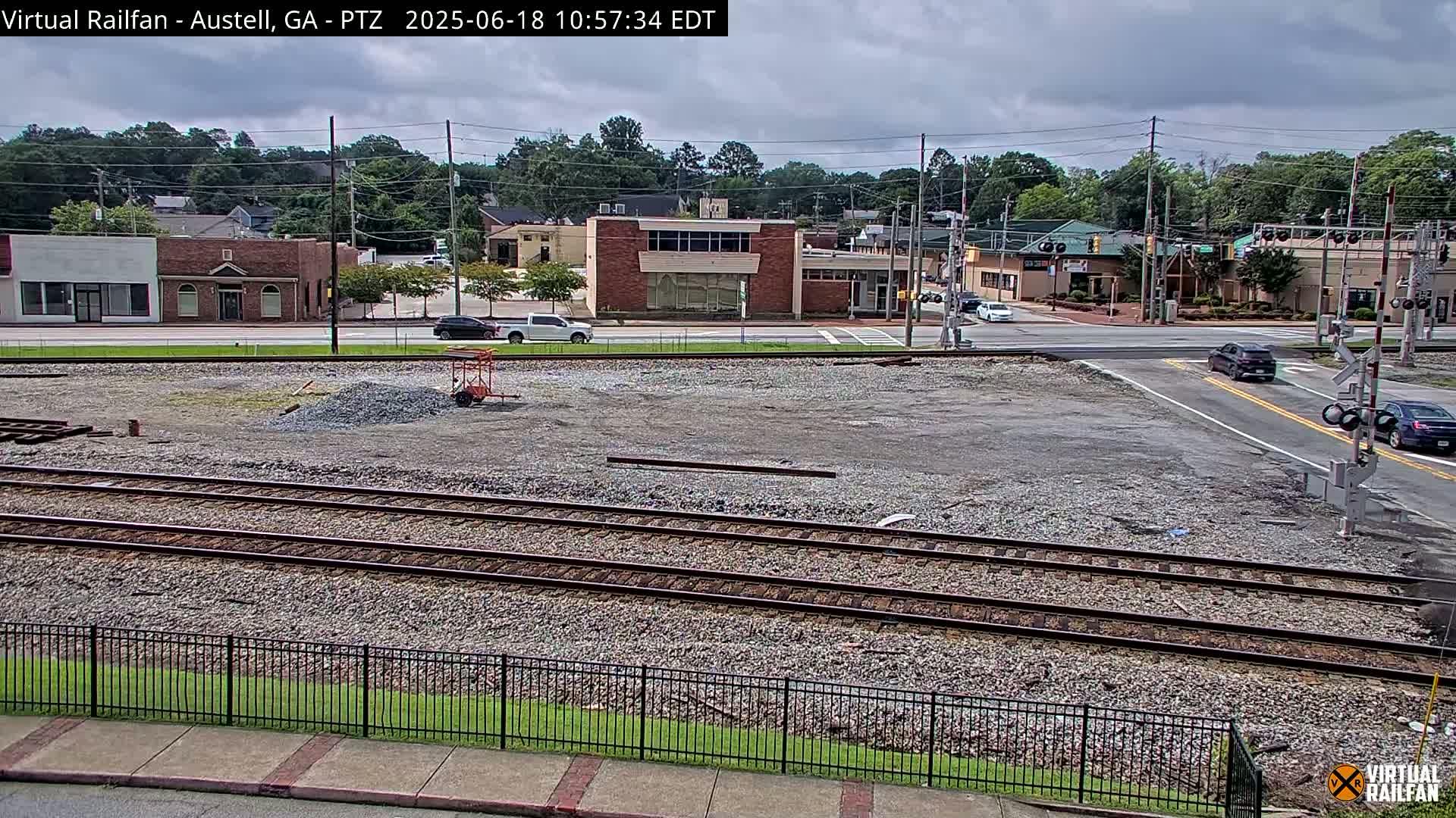 A mostly cloudy day reveals a gravel lot with train tracks in front of a street with several buildings, some cars, and railroad crossing signals.