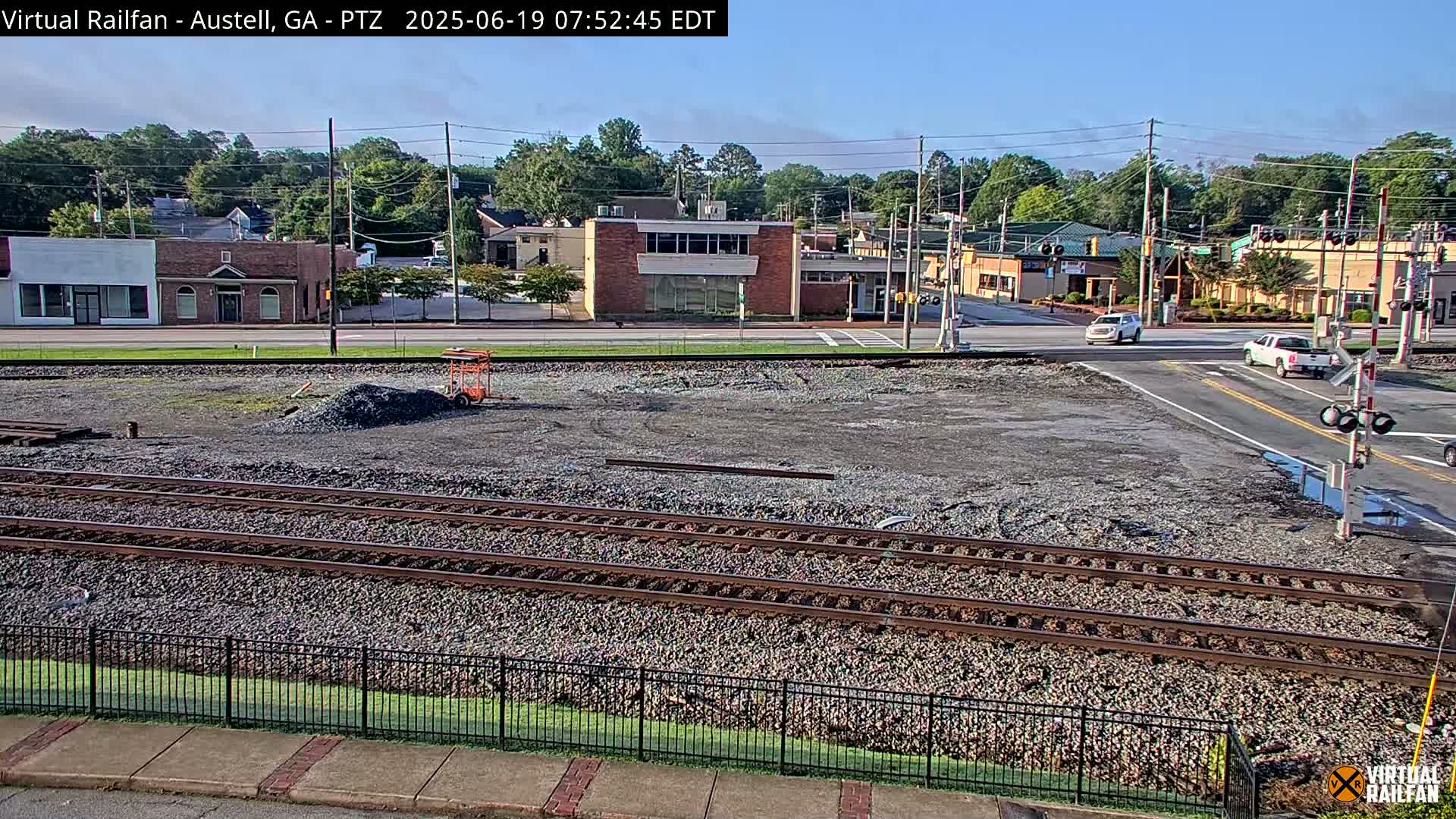 A mostly clear day reveals a view of railroad tracks, a gravel lot, and a street intersection with buildings and cars visible in the background.