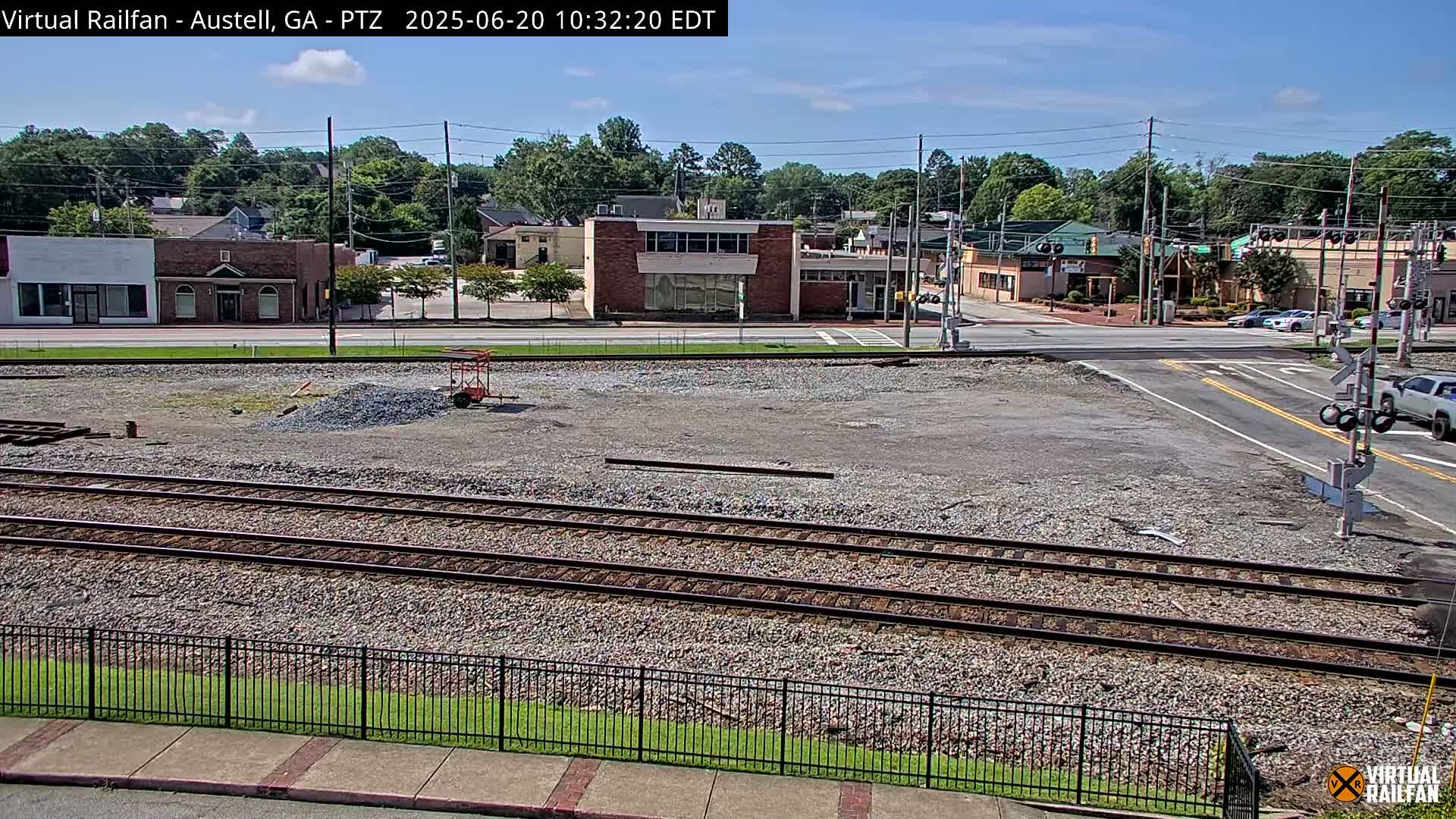 A mostly sunny day shows railroad tracks in a gravel lot next to a street intersection with several buildings visible in the background.
