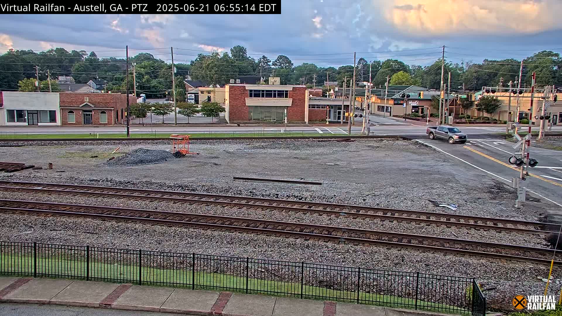 A mostly clear evening sky hangs over a railroad crossing near a street intersection with several buildings and a pickup truck driving across.