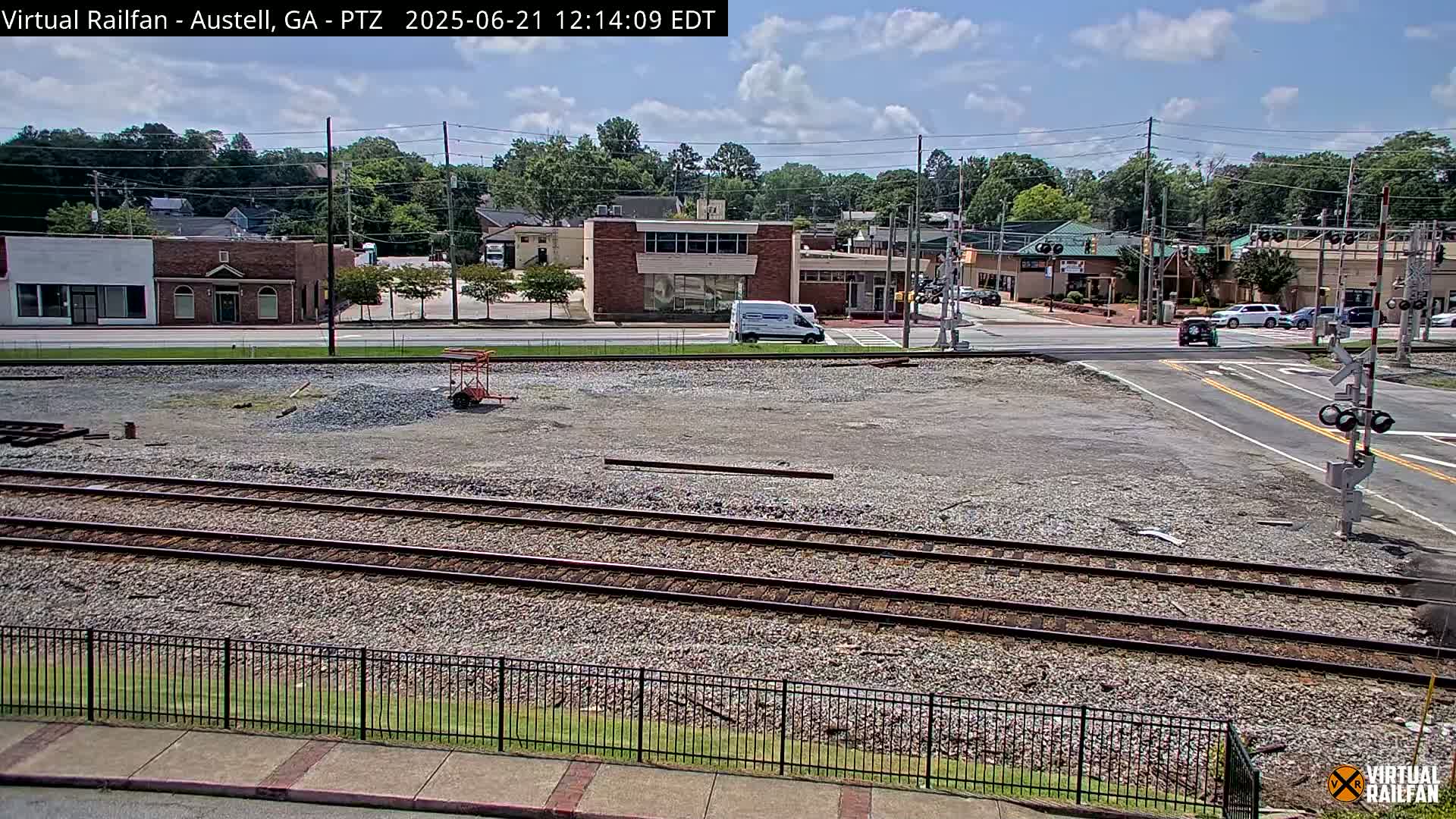 A mostly sunny day shows a view of railroad tracks next to a street with buildings and cars, a large empty lot, and some construction equipment visible.