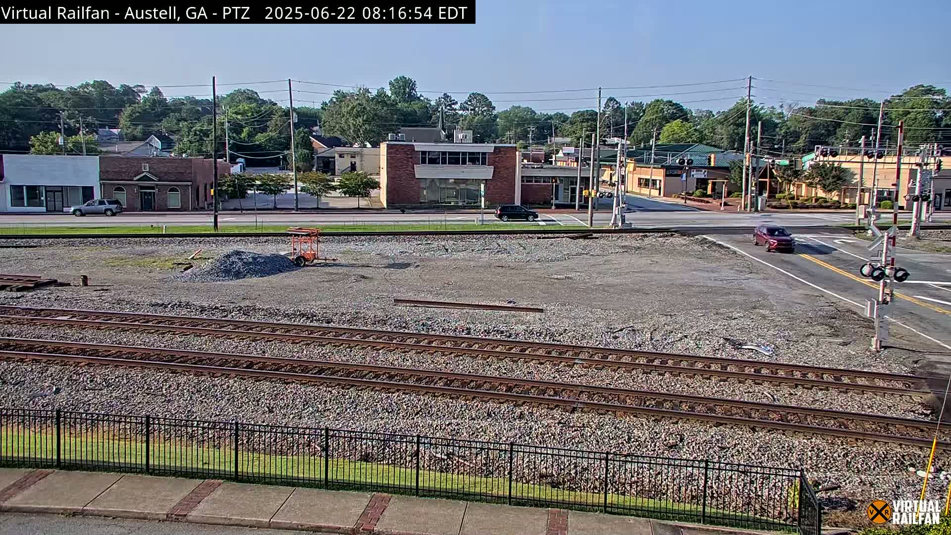 A mostly sunny view of a railroad crossing near a town, with several buildings visible in the background and a pile of gravel in the foreground.