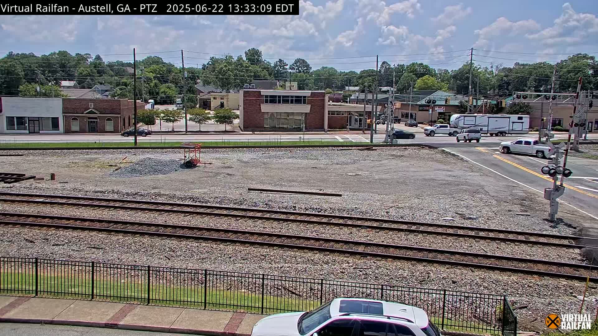 A sunny day shows railroad tracks in a gravel area next to a street with buildings and cars, including an RV, under a partly cloudy sky.