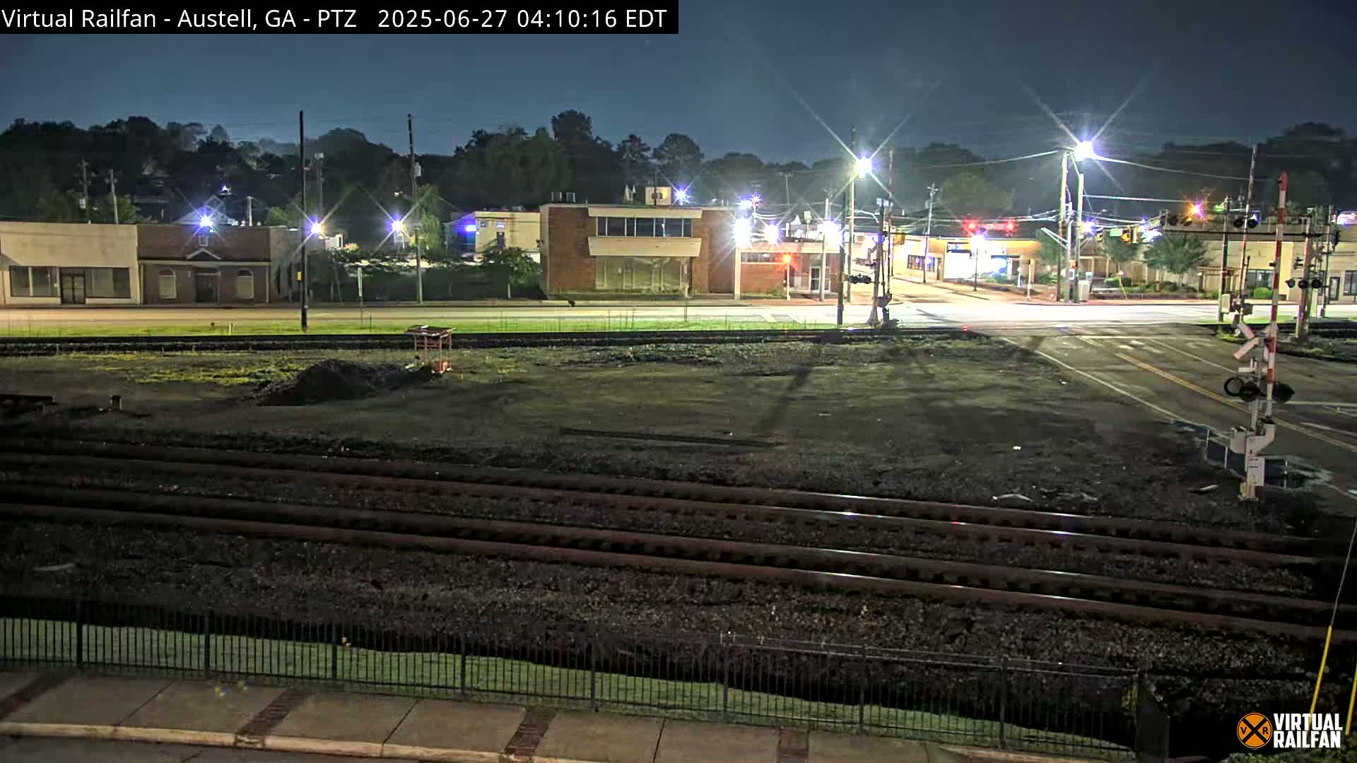 A nighttime view of a railroad crossing near a street intersection, with several buildings visible in the background under clear skies.