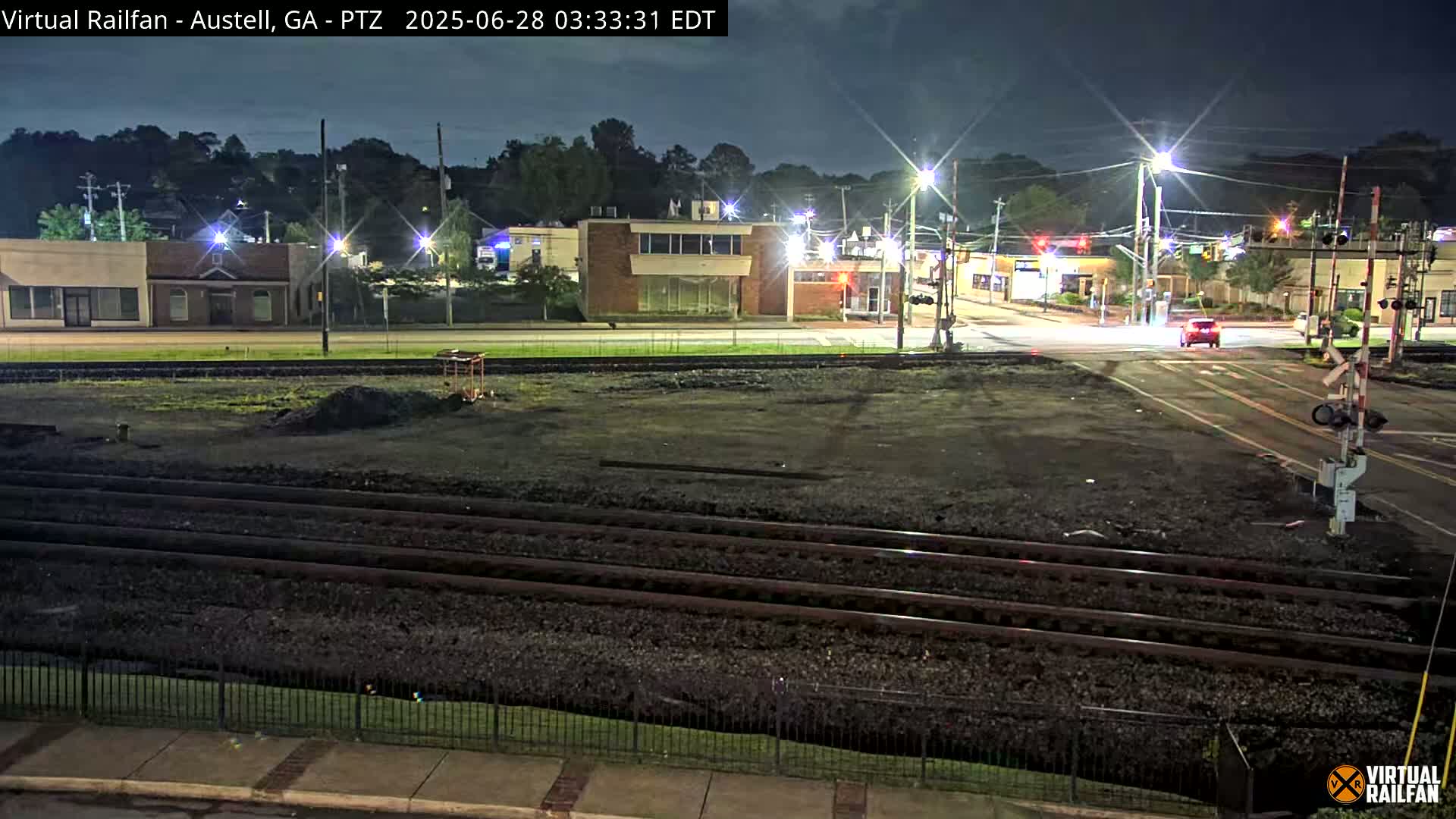 A nighttime view of a railroad crossing near a street intersection with several buildings in the background under a dark sky.
