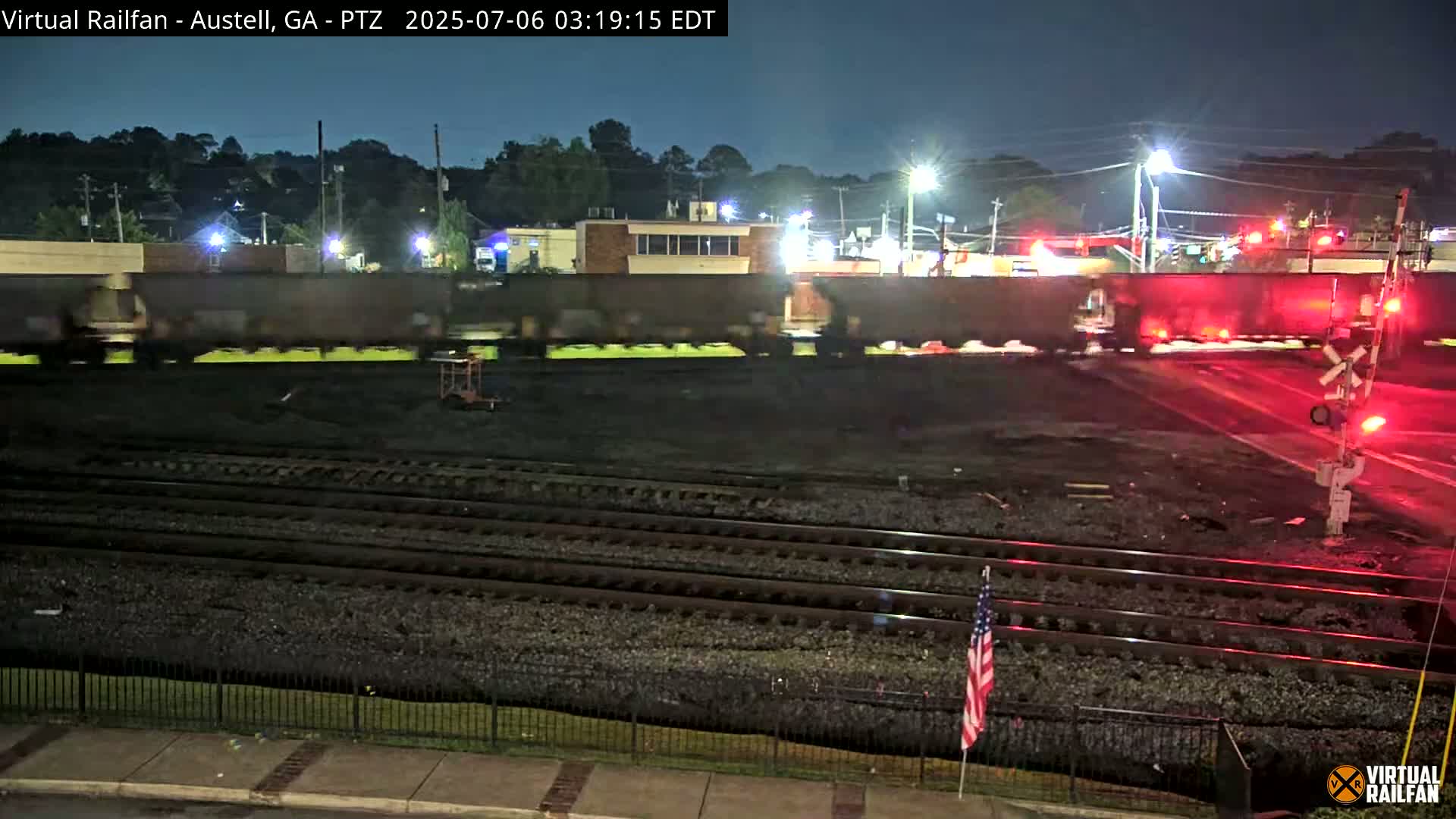 A long freight train passes through a nighttime railroad crossing under illuminated streetlights.