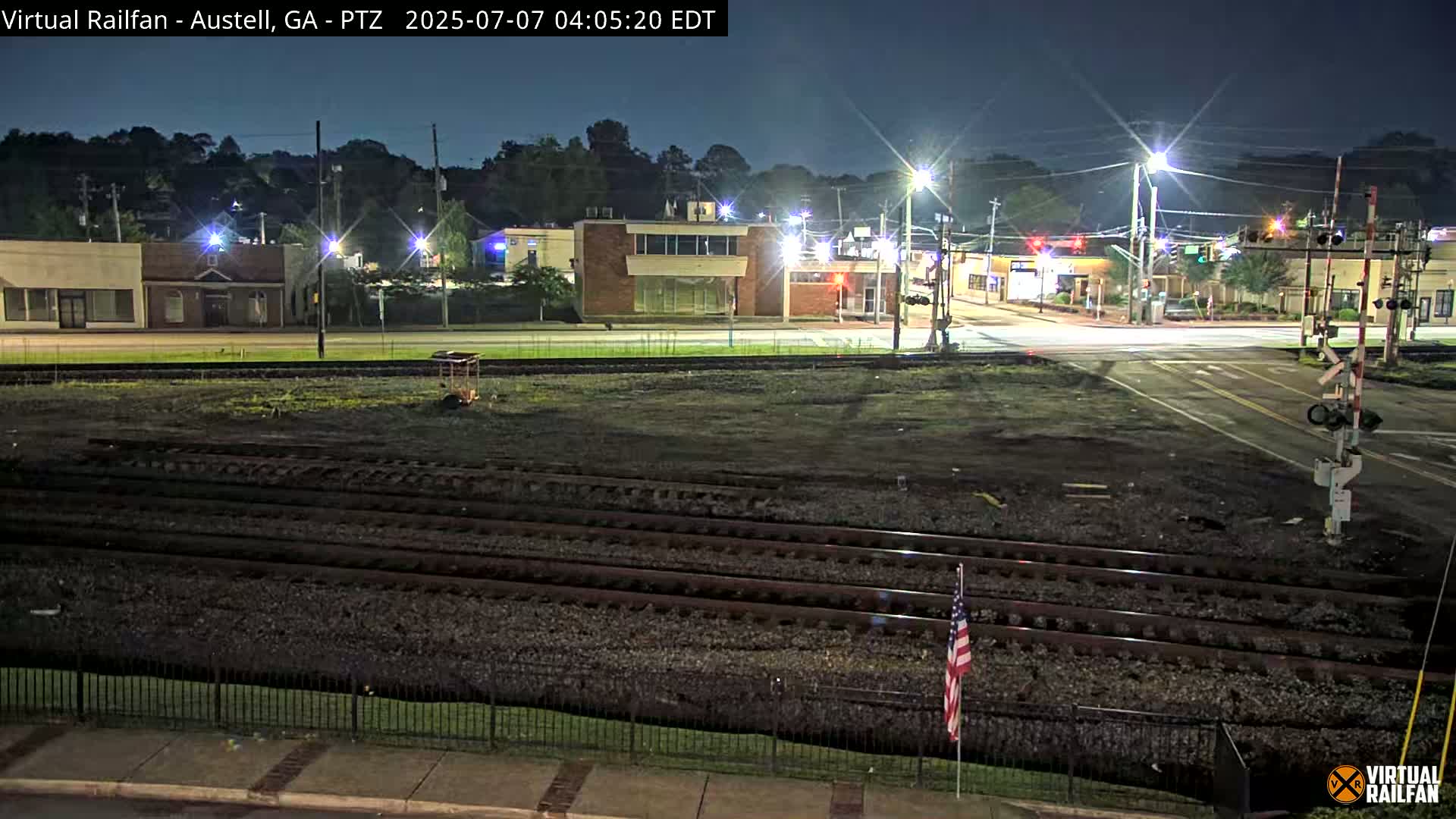 At night, a mostly empty intersection with a railroad crossing and several buildings is illuminated by streetlights.