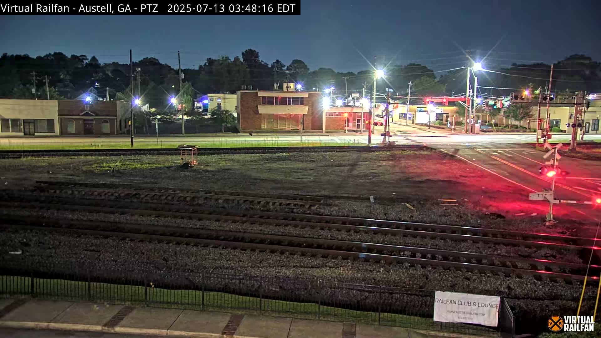 A nighttime view of a railroad crossing with train tracks in the foreground, and a street intersection with buildings and streetlights in the background under a clear, dark sky.