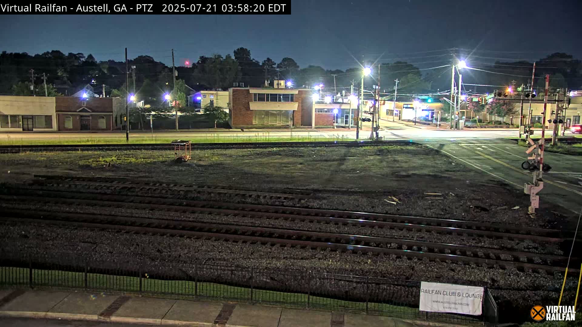 At night, a mostly empty train yard is adjacent to a street intersection with several buildings under streetlights.