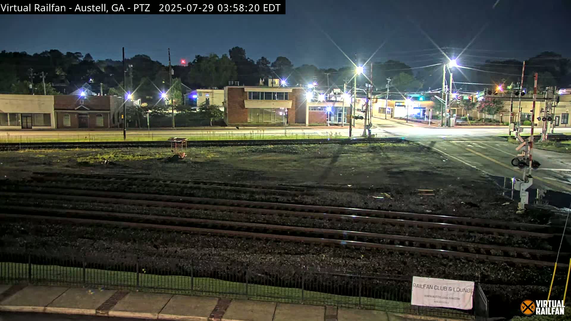 A nighttime view of railroad tracks in front of a street intersection with several buildings under a clear sky.