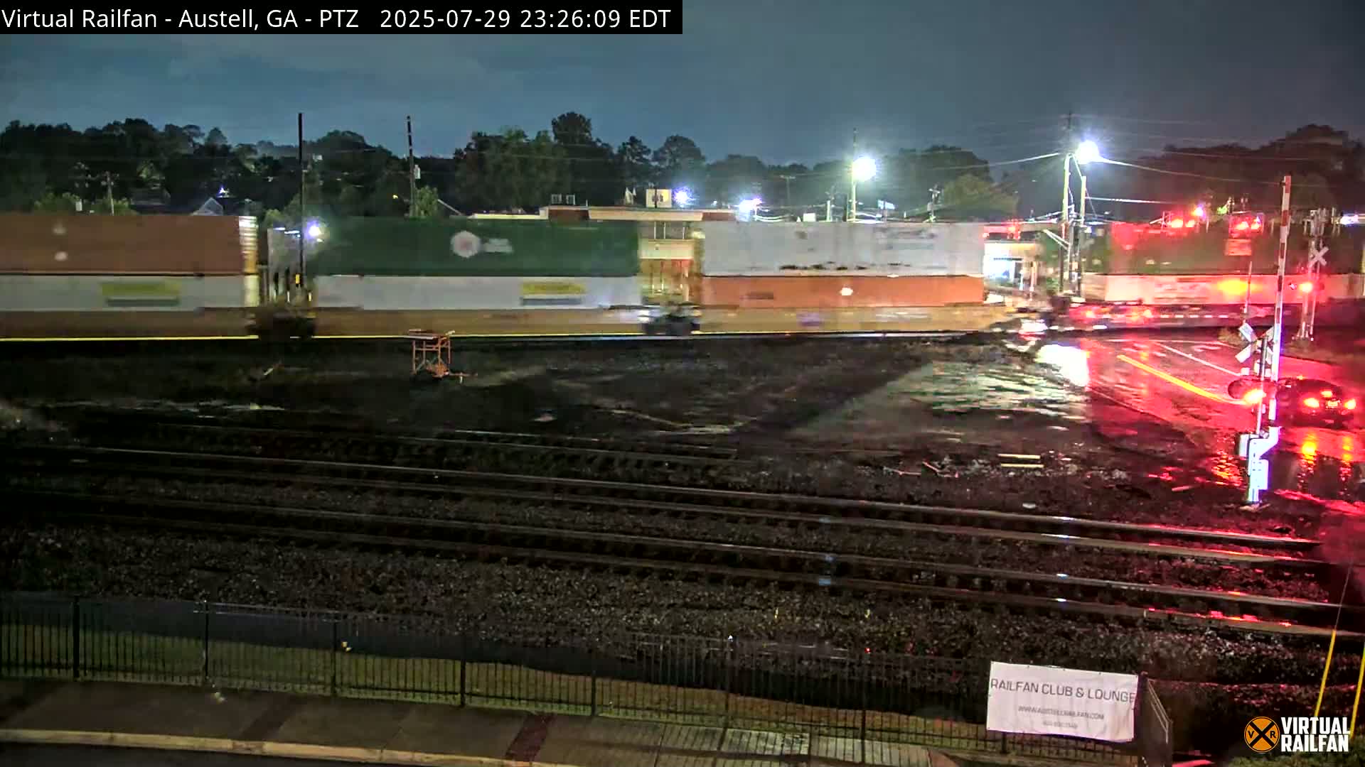 A nighttime view of railroad tracks in front of a street intersection with several buildings under a clear sky.