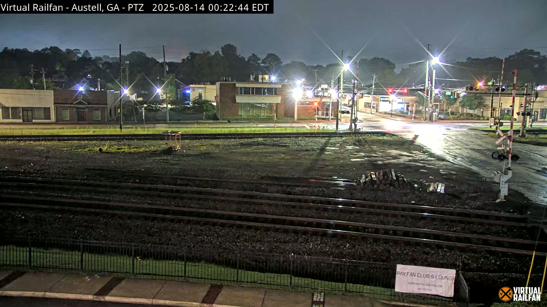 A nighttime view of railroad tracks in front of a street intersection with several buildings under a clear sky.