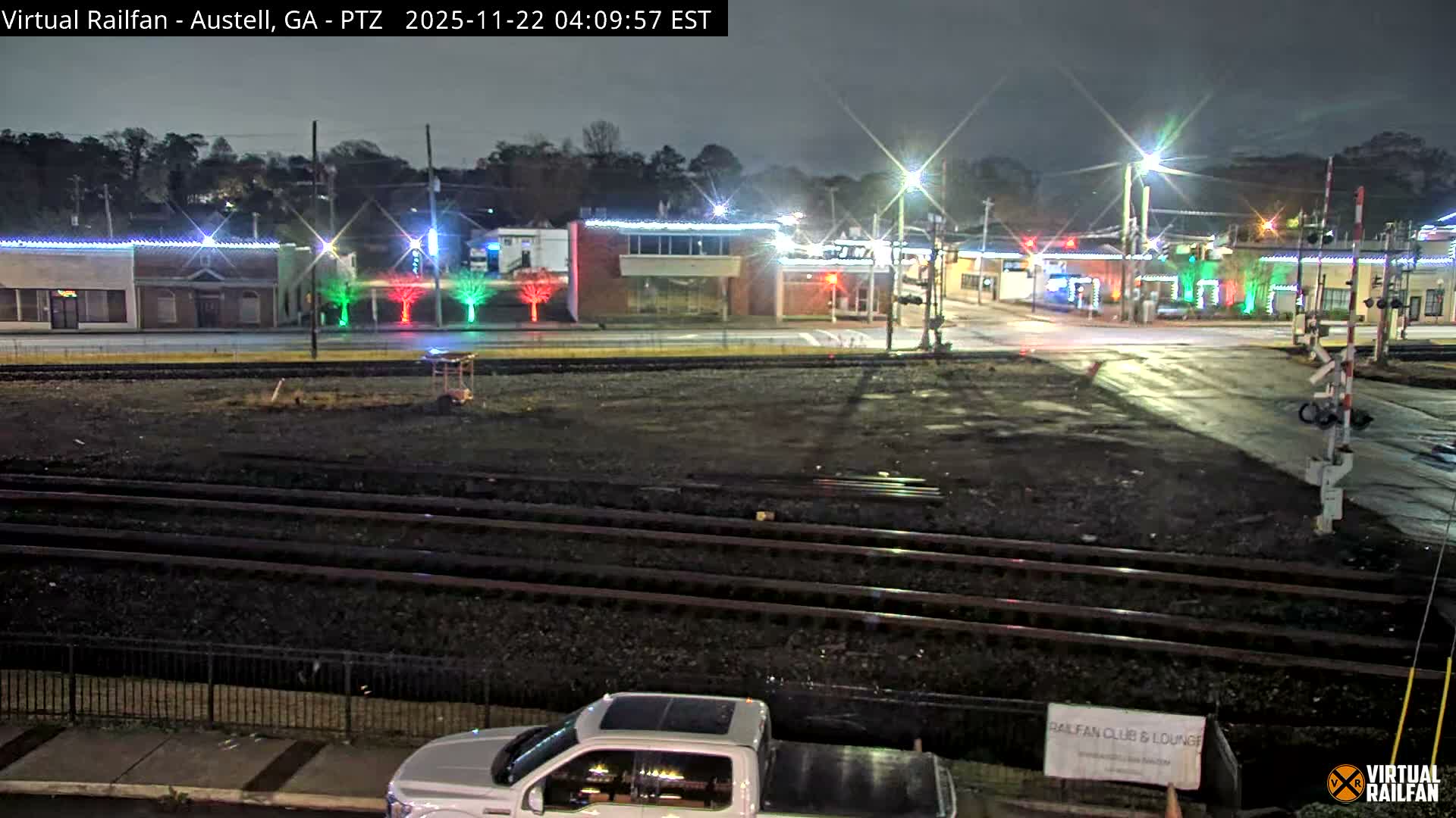 An overcast night reveals a multi-track railroad crossing through a town, where buildings are adorned with bright festive red and green lights, and the wet ground reflects the streetlights.