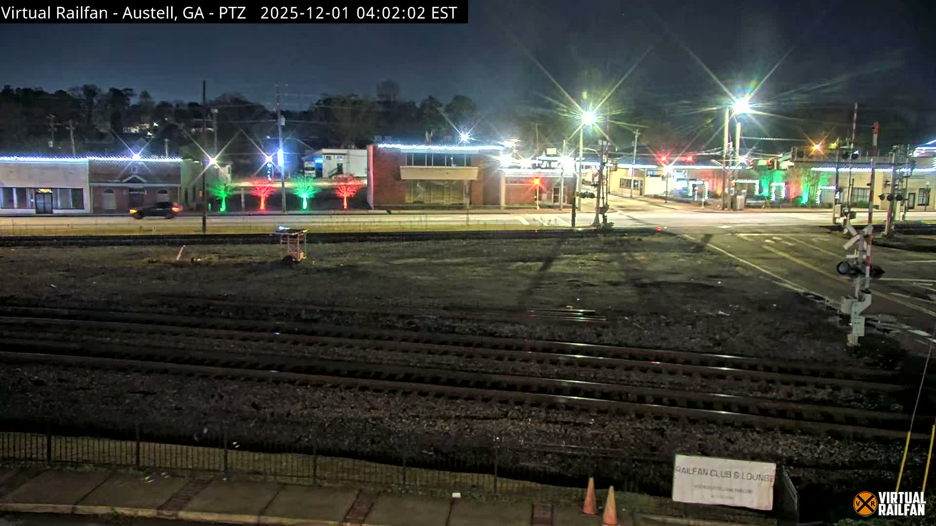 A clear night view reveals multiple railroad tracks in the foreground, leading to a street lined with brightly lit buildings, streetlights, a moving car, and trees decorated with festive red and green lights.